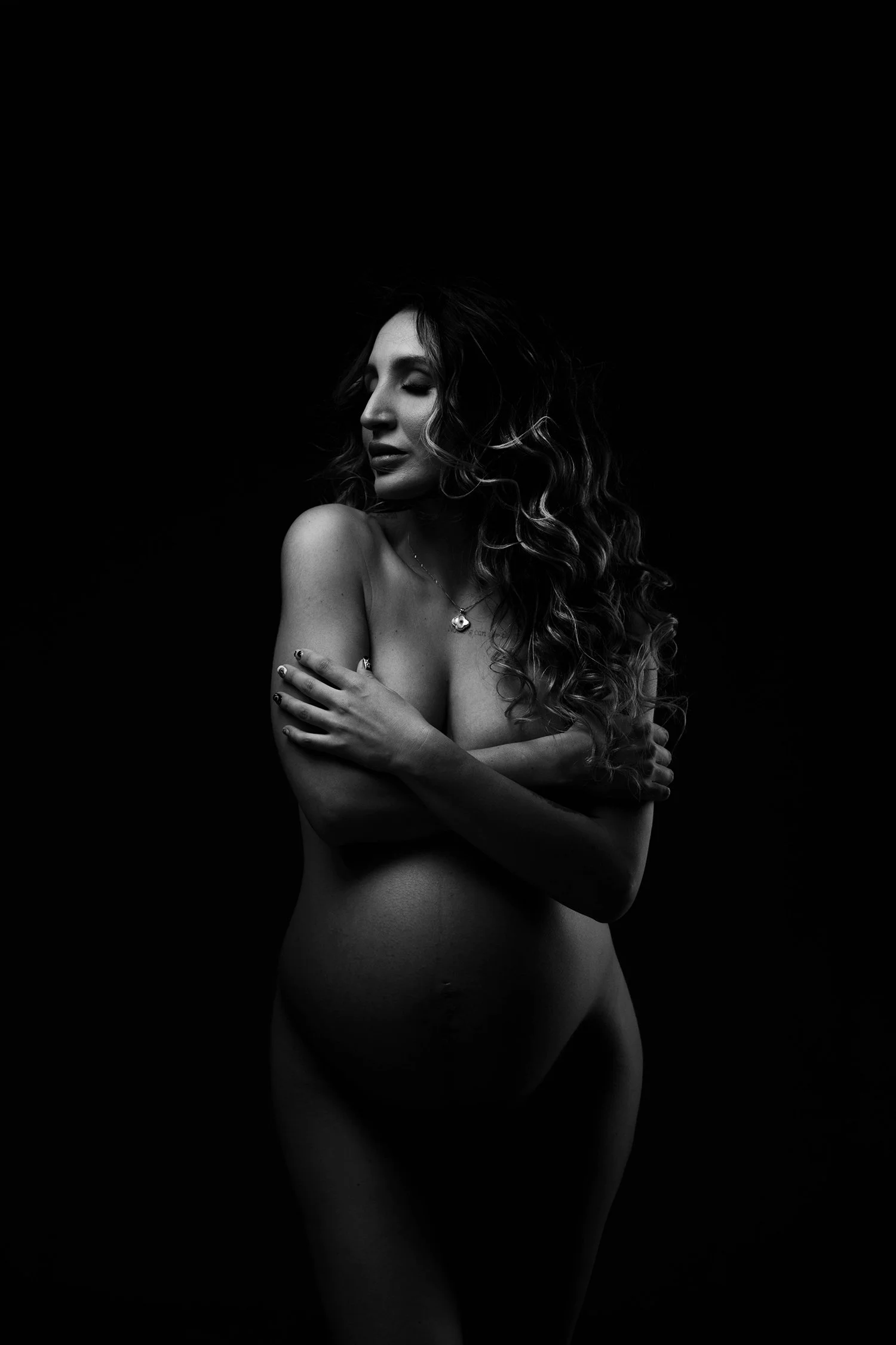Black and white photo of a topless woman with curly hair, posed with arms crossed over her chest, against a dark background.