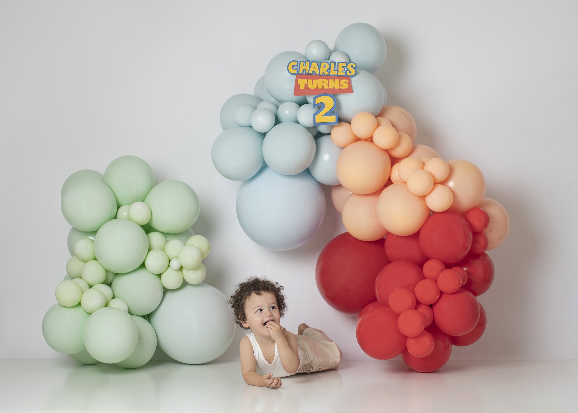 A young child with curly hair lying on the floor smiling and holding their finger to their mouth, in front of a backdrop of large pastel-colored balloons arranged in clusters, with a sign that reads "Charles Turns 2".