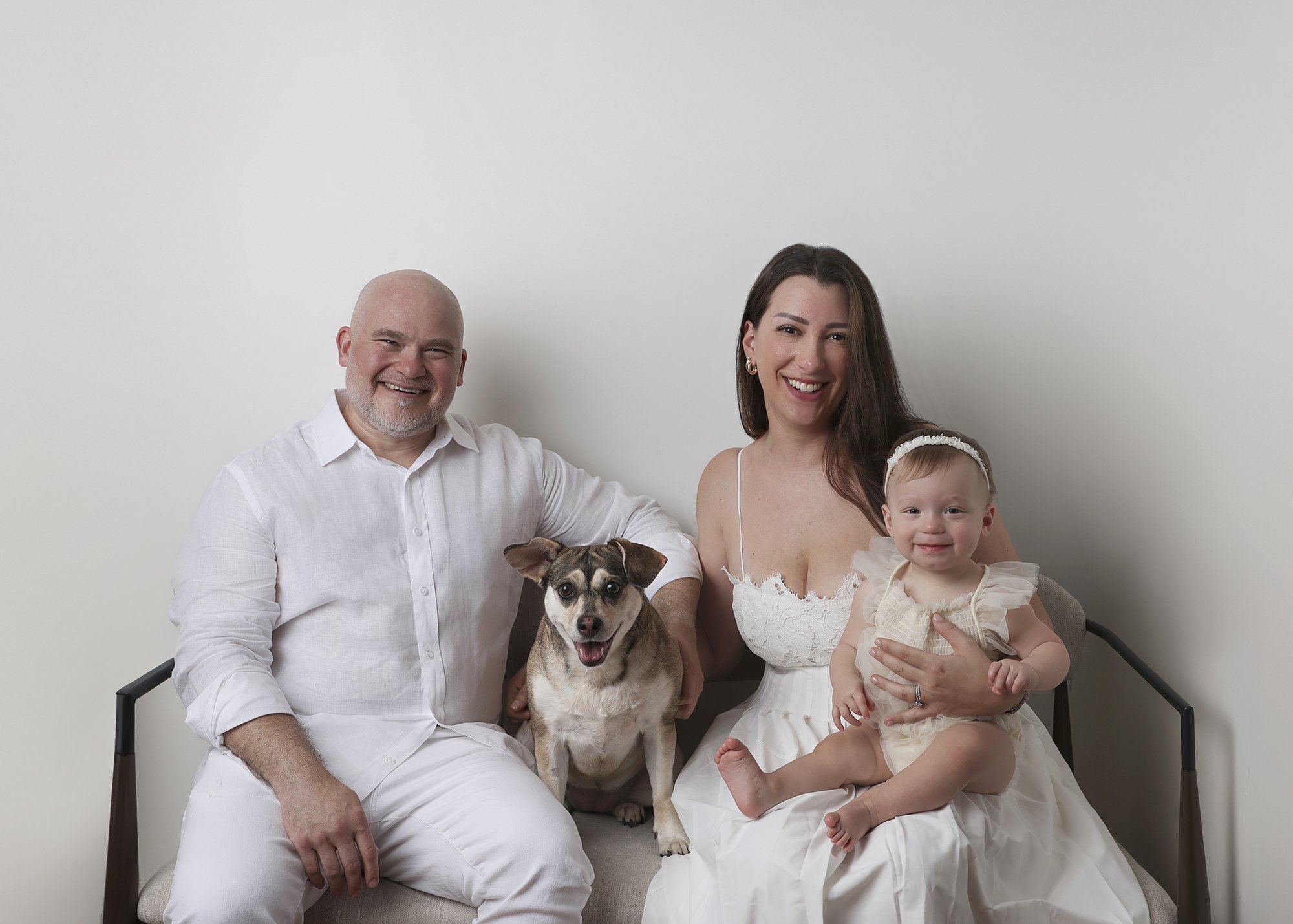 A happy family of three with a dog and a baby girl sitting on a white chair against a plain light wall. The man has a white shirt, the woman has a white dress, and the girl is in a light-colored dress with a headband.