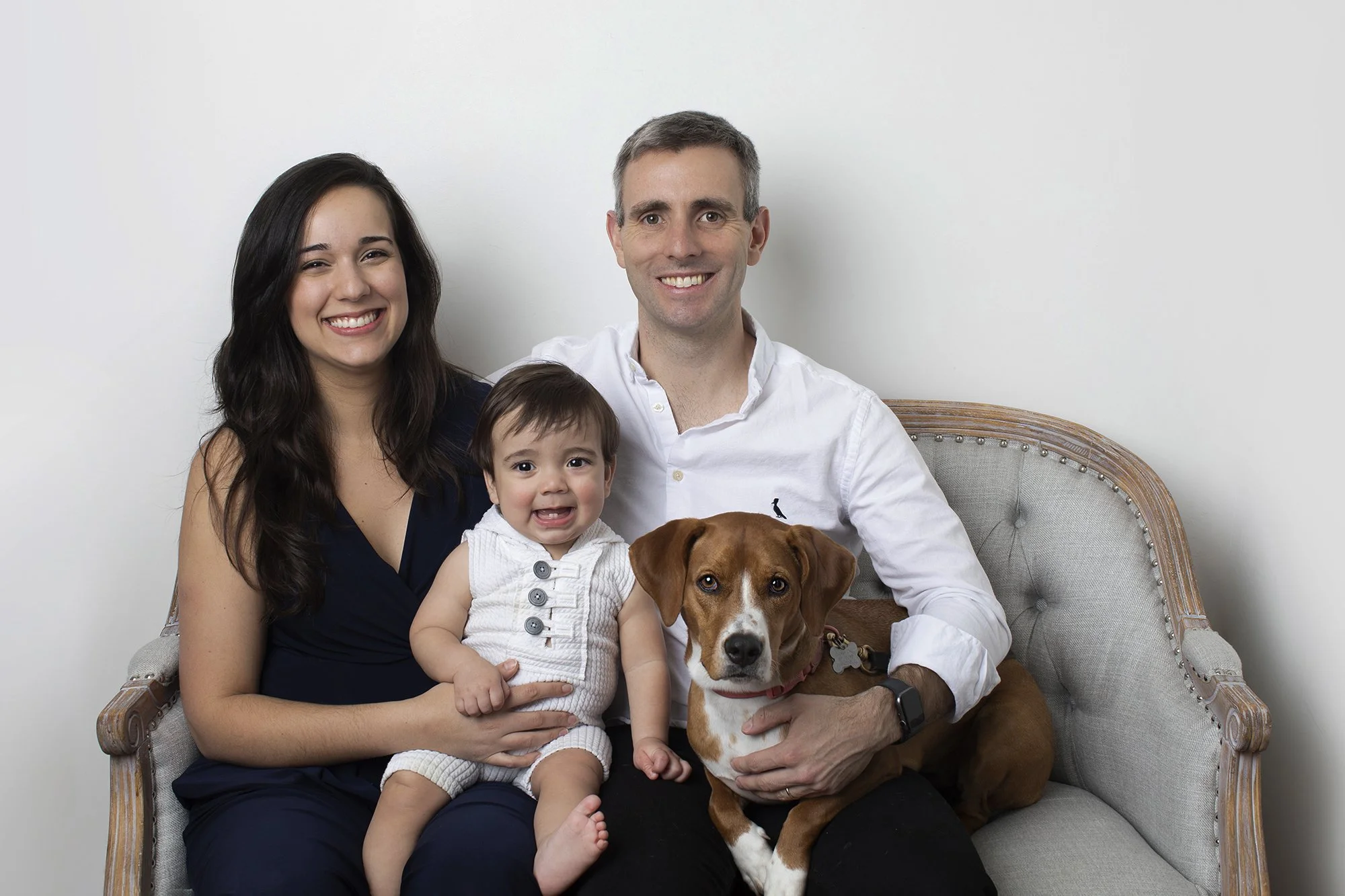 A happy family of four, including a woman, a man, a young girl, a brown and white dog, and a small child, all sitting together on a vintage-style sofa against a plain white wall.