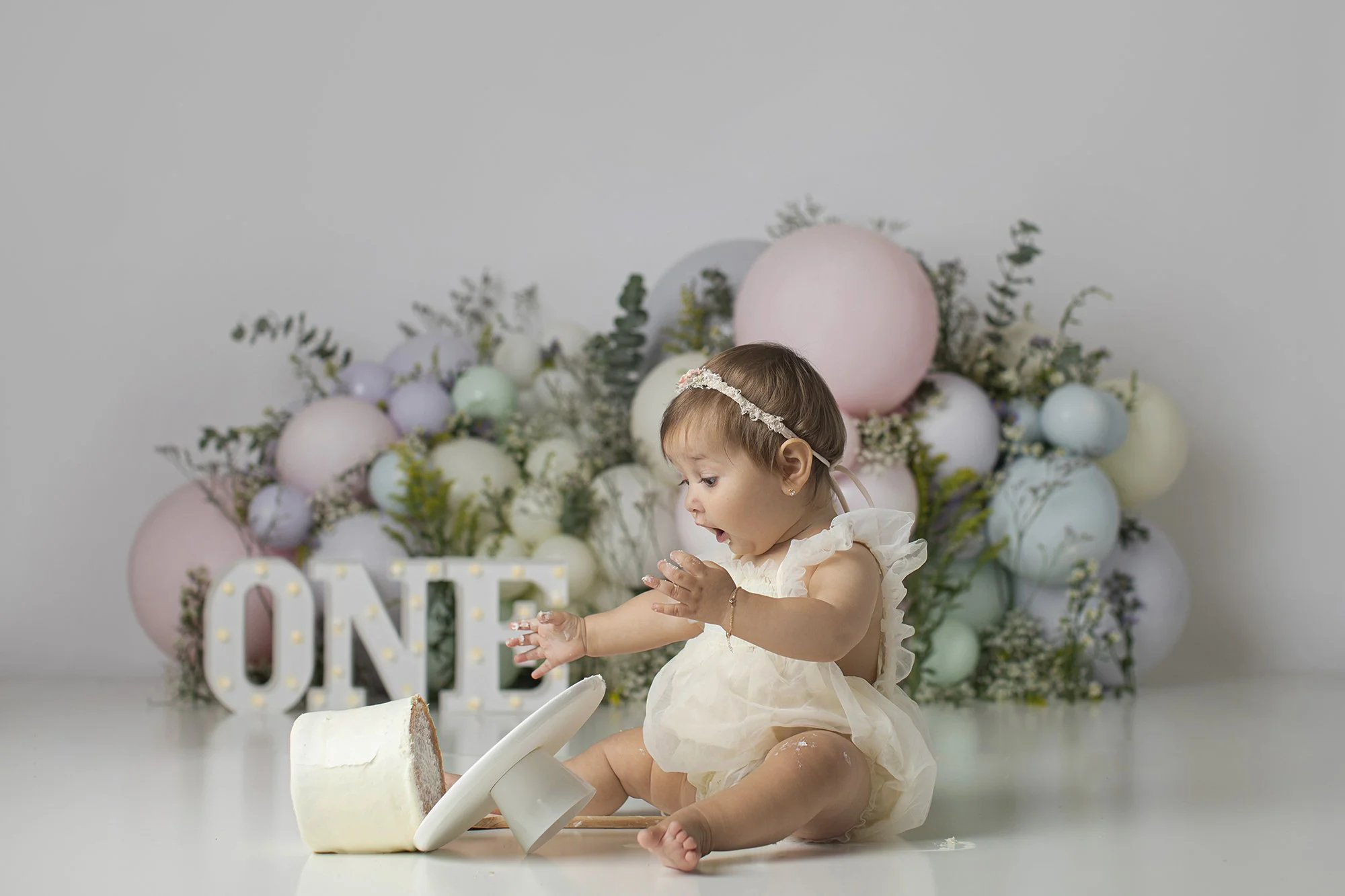 Young girl with a headband in a cream dress playing with a cake and cake stand on a white surface in front of pastel balloons and floral decorations, with a white 'LOVE' sign in the background.