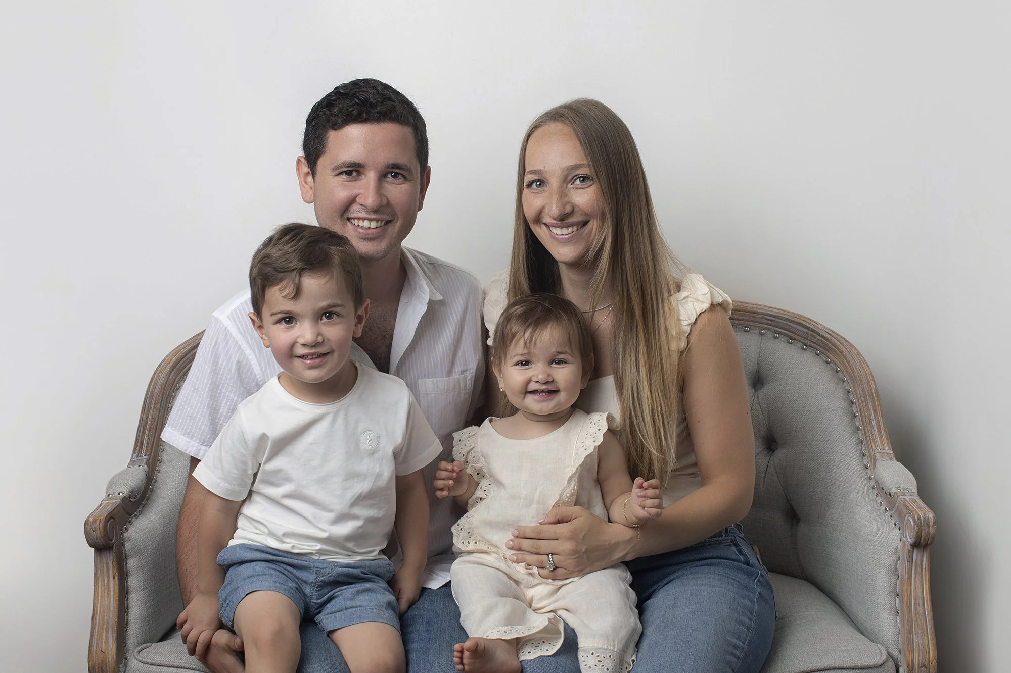 A family of five sitting on a vintage-style sofa against a plain white wall, smiling at the camera.