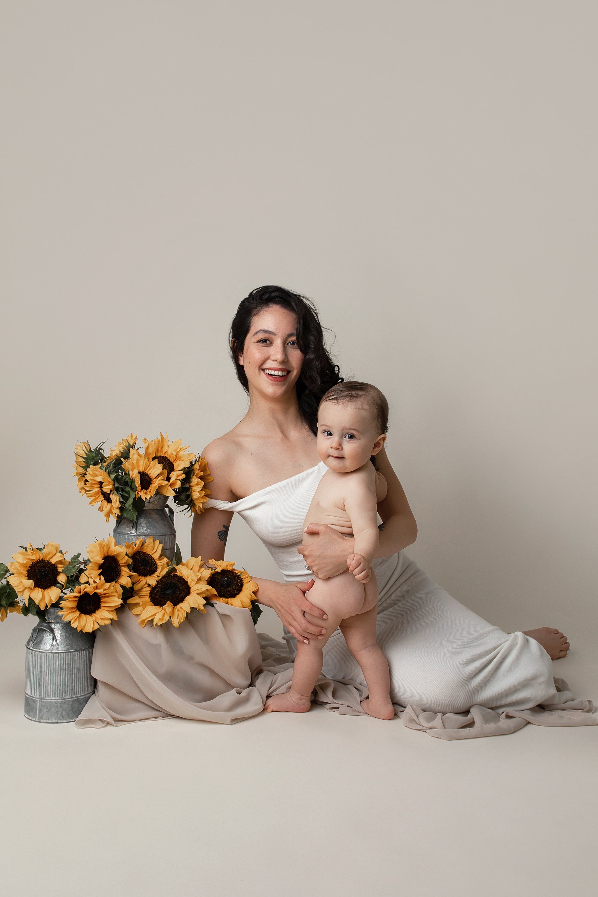 A smiling woman with dark curly hair holding a baby, seated on the floor next to a large bouquet of sunflowers in vases, against a neutral background.