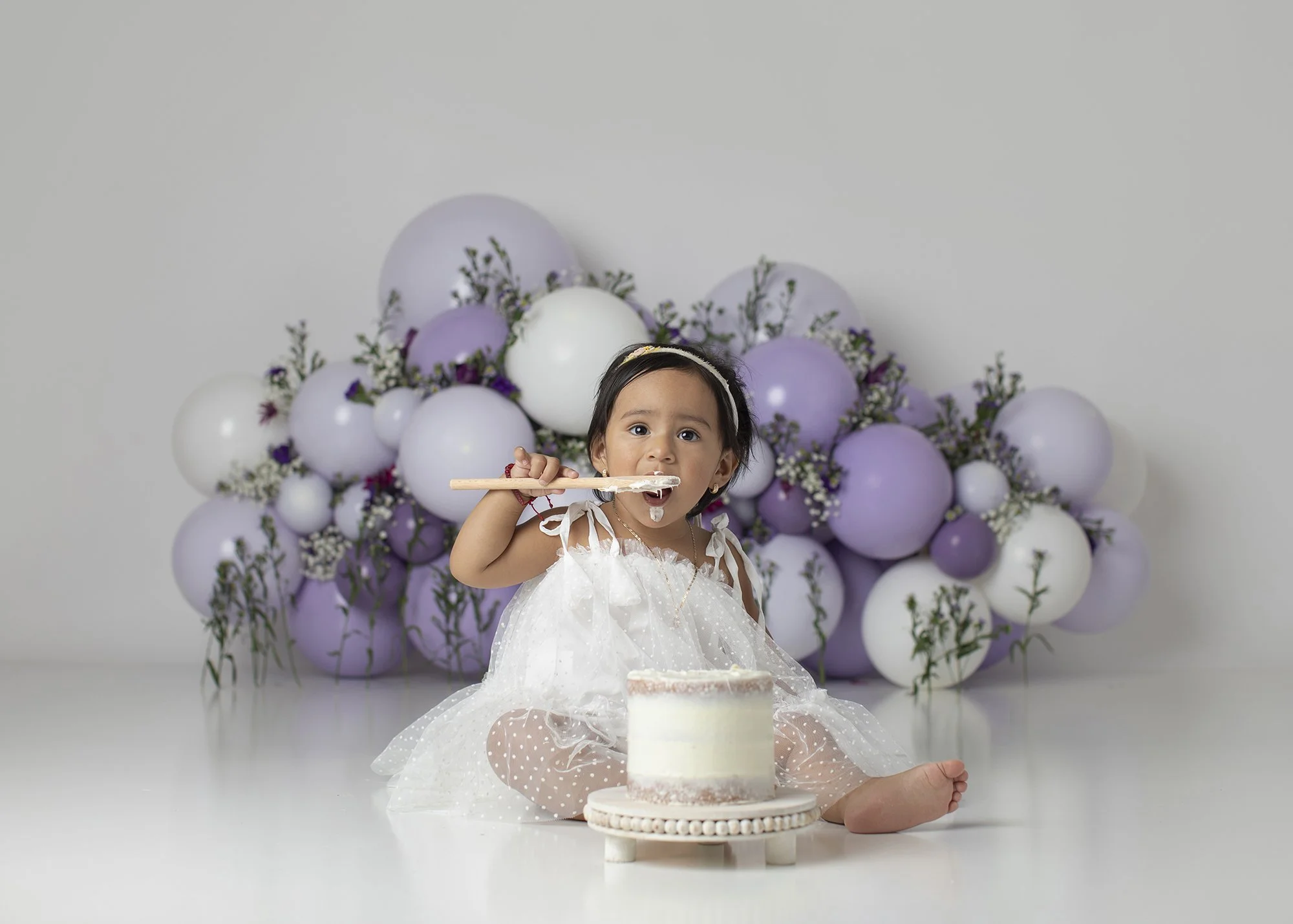 A young girl in a white dress with polka dots and a headband, sitting on the floor, holding a spoon and about to eat a small white cake with pink accents. Behind her is a floral backdrop with white and purple balloons and flowers.