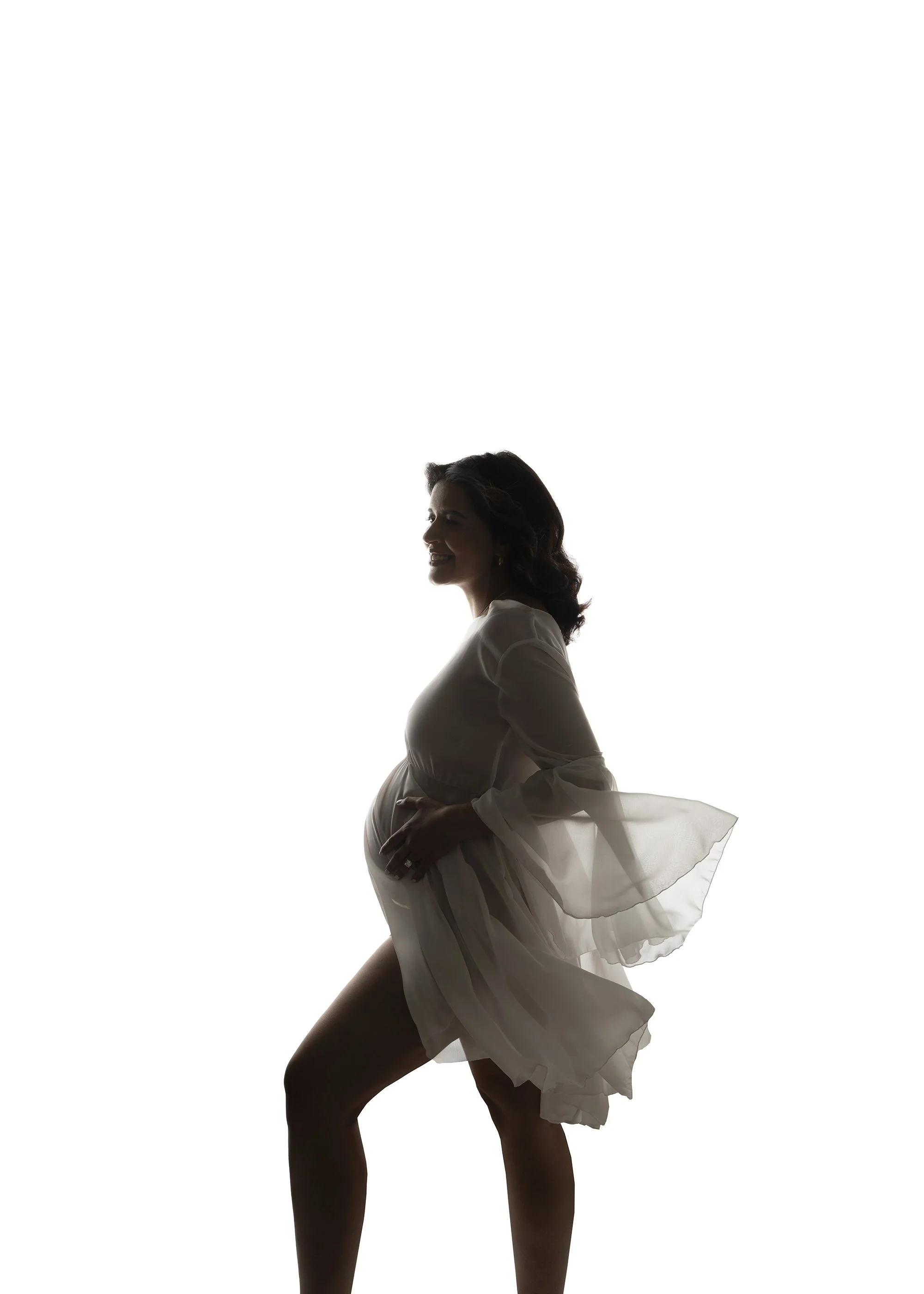 Silhouette of a pregnant woman in a flowing dress, smiling, with her hand on her belly, against a bright light background.