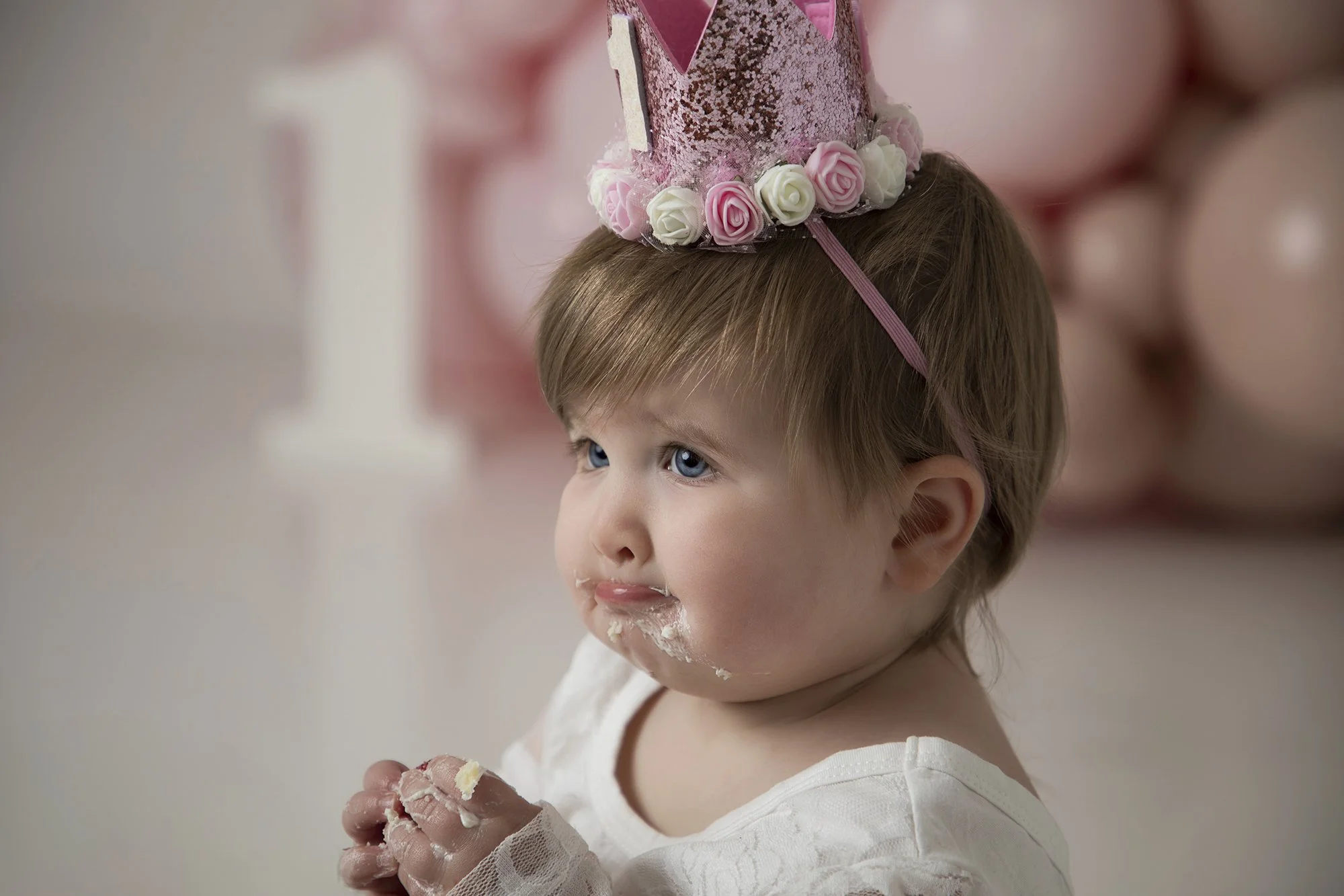 A young girl with blue eyes and light brown hair, wearing a white dress and a pink birthday hat decorated with roses and glitter, is holding a piece of cake with white frosting, and has some frosting on her face and hands.