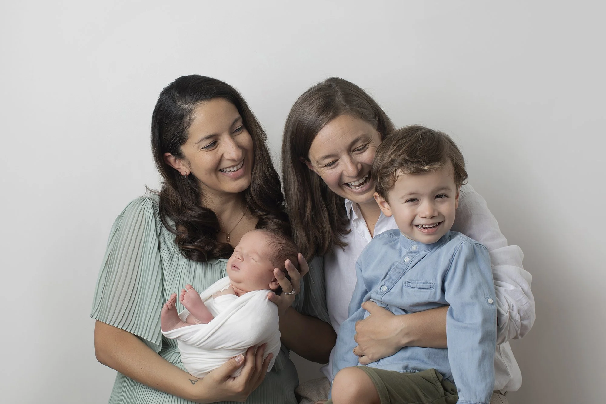 Two women and two young children, a boy and a girl, smiling and cuddling against a plain gray background.