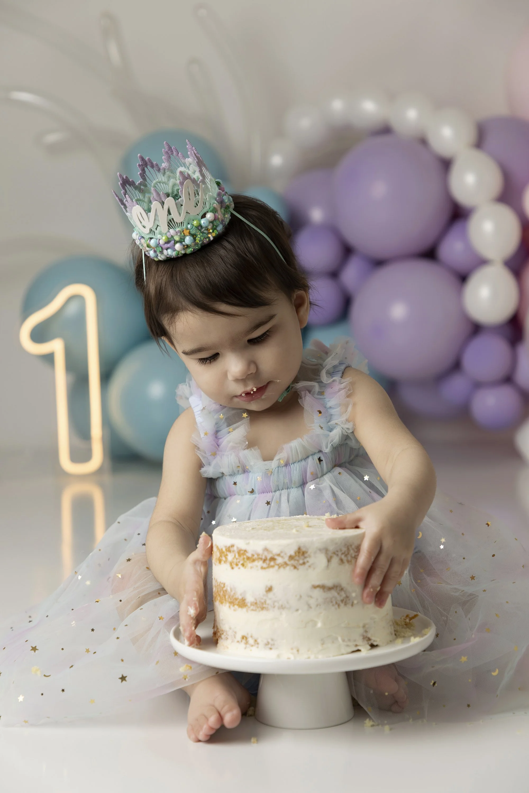 Young girl wearing a pastel-colored dress with star patterns and a birthday crown, sitting in front of a birthday cake, surrounded by purple and white balloons, celebrating her birthday.
