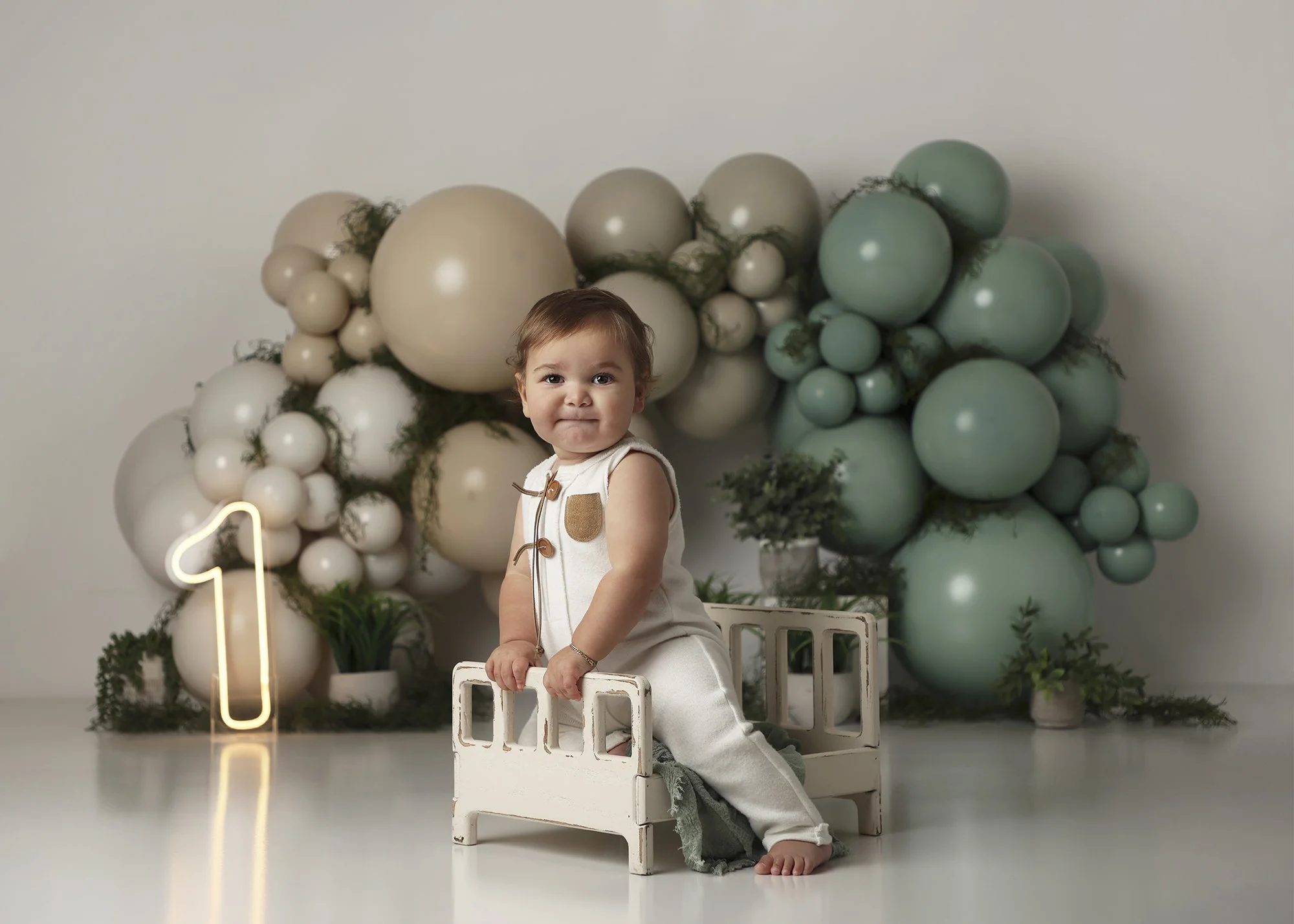 A young child with brown hair sitting on a small white wooden bench, surrounded by a backdrop of beige, white, and mint green balloons, with greenery and a neon number one decoration in the scene.