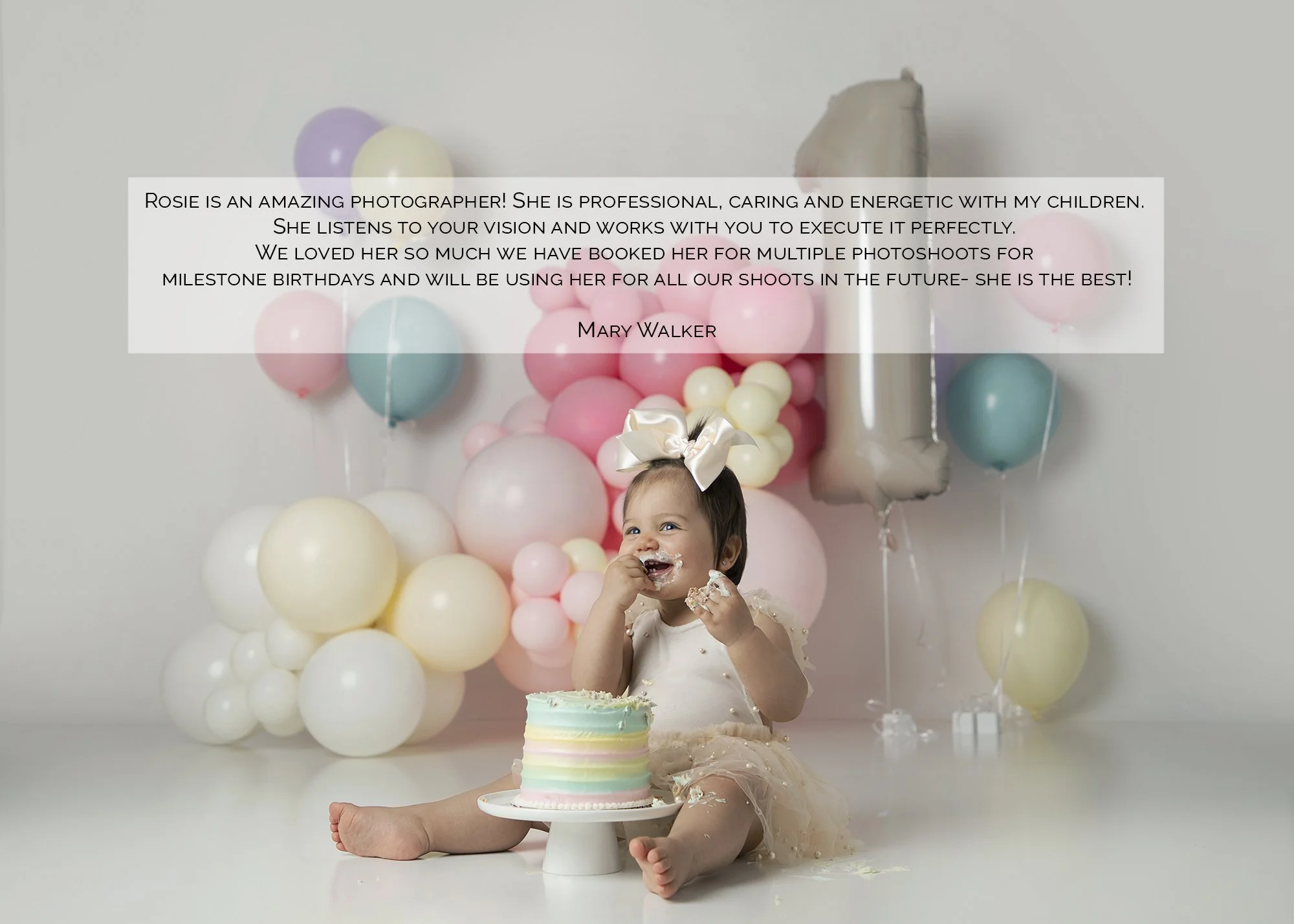 A young girl sitting on the floor celebrating her birthday with a colorful cake, balloons, and a birthday hat, with a gray wall background.
