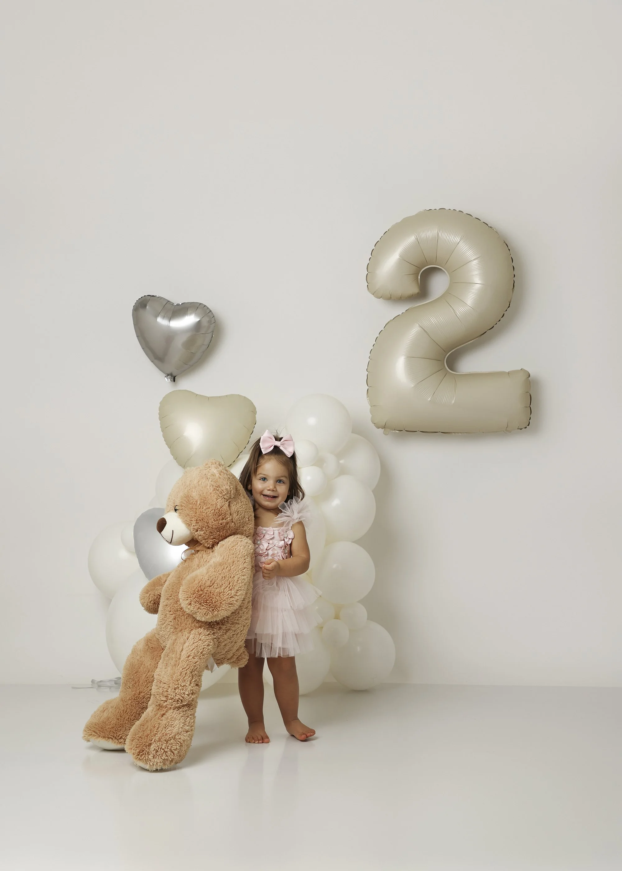 A young girl in a pink dress holding a large teddy bear, standing in front of white balloons and silver number balloons forming the number 2, with a small pink bow in her hair, smiling inside a room with a plain white wall.