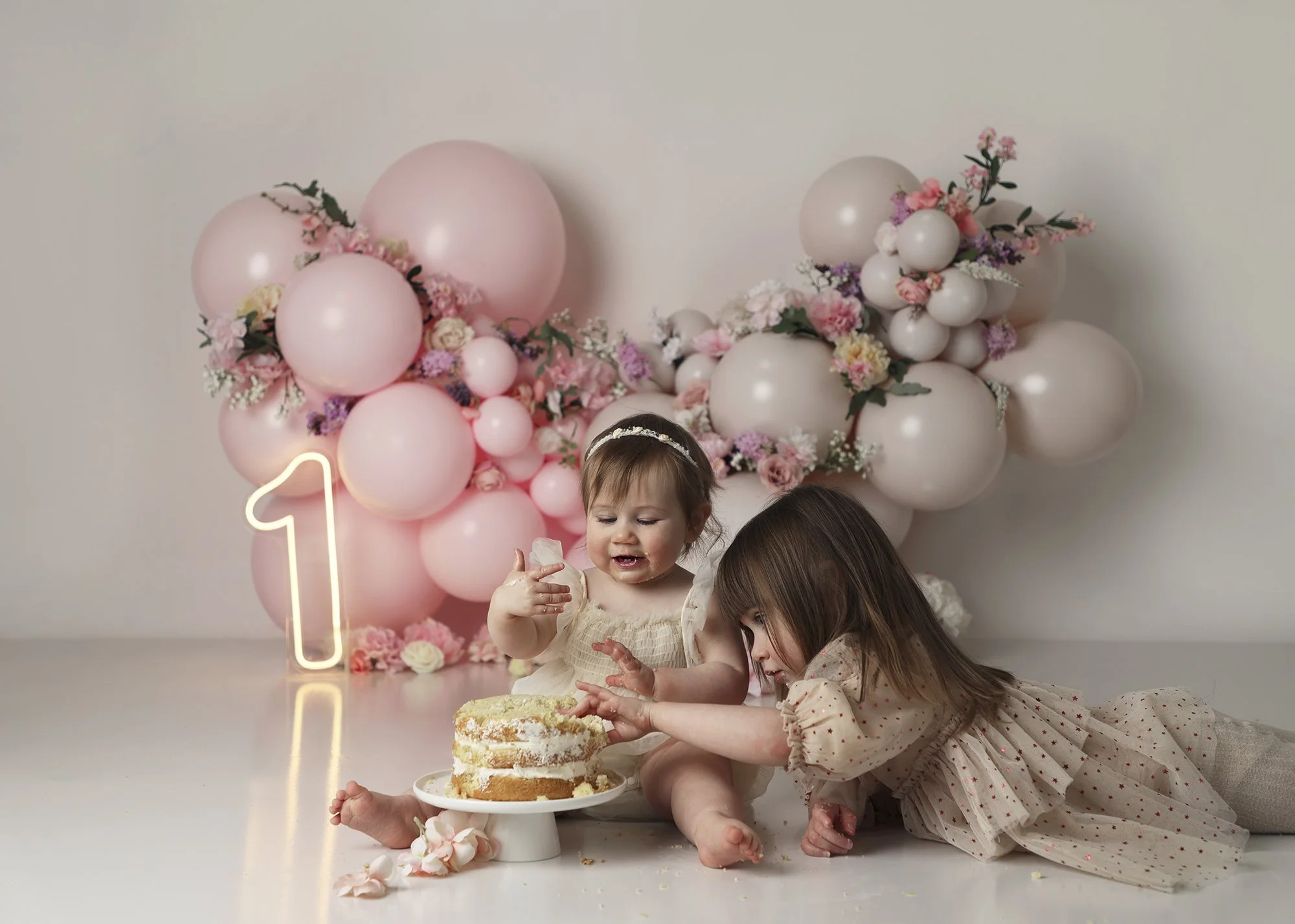 Three children celebrating a birthday with pink balloons and flowers in the background, a cake and a neon number one sign on a white table, one girl is reaching for the cake.