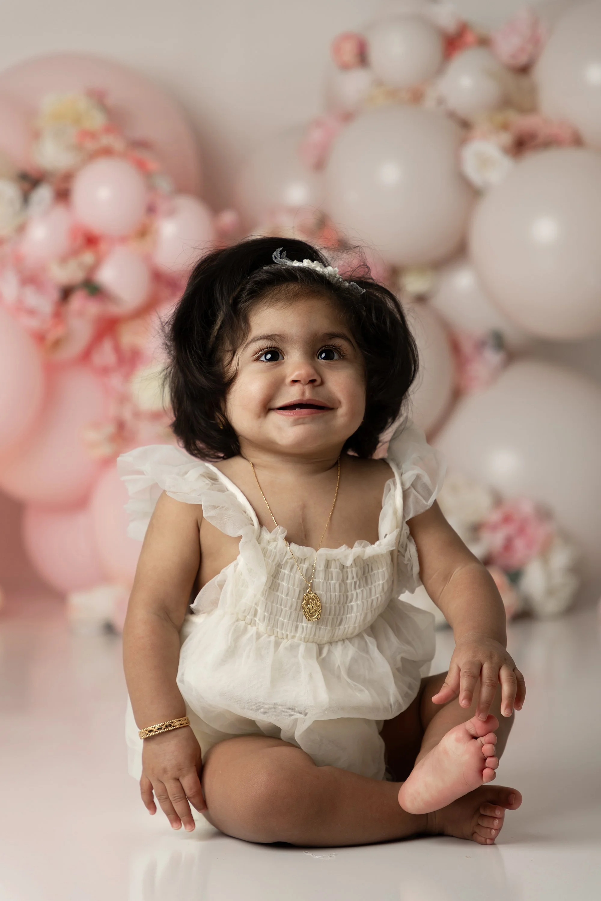 A young girl with dark curly hair wearing a cream-colored dress with puffed sleeves, seated on a person's lap, with a backdrop of pink and white balloons decorated with flowers. The girl is smiling and looking slightly upward.