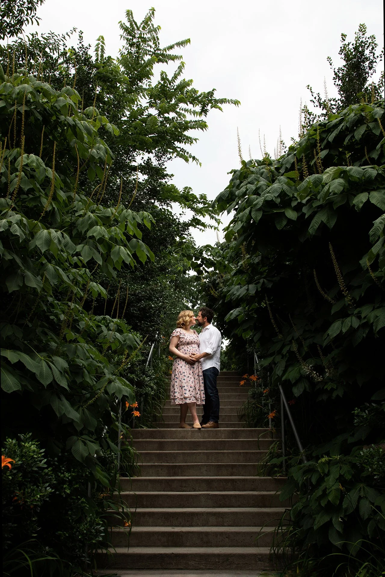A couple stands on a staircase surrounded by lush green foliage, sharing a romantic moment and looking into each other's eyes.