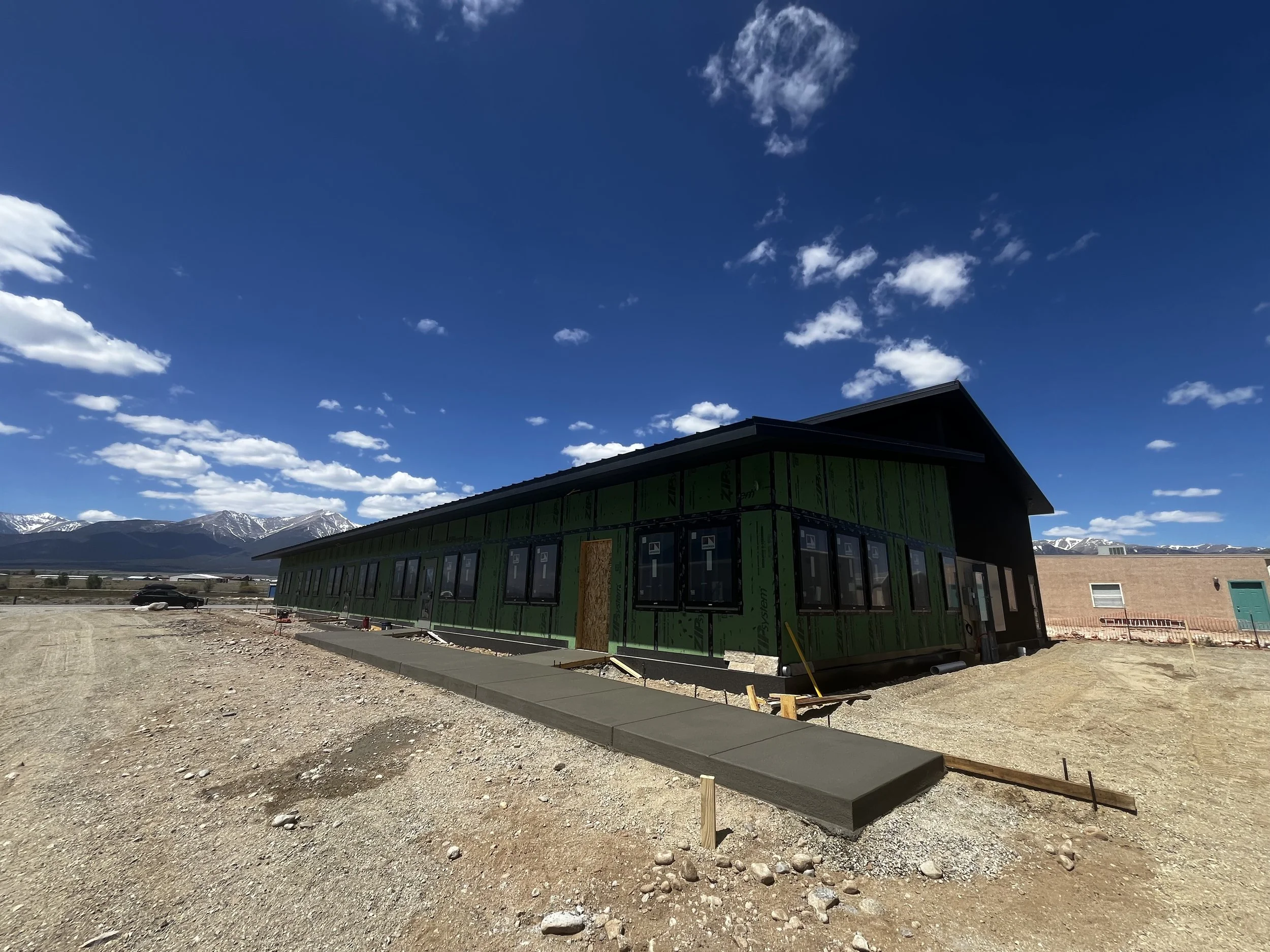 Under construction house with green sheathing, concrete sidewalk, dirt lot, mountains in background, blue sky with clouds.