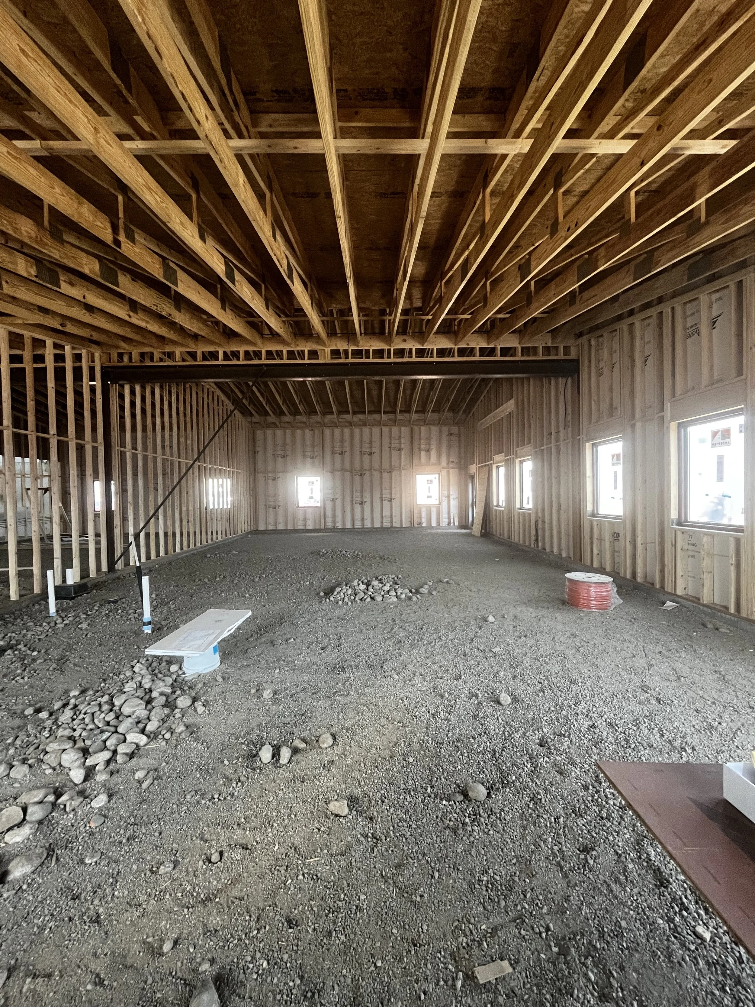 Interior of a building under construction, showing exposed wooden framing for walls and ceiling, with several small windows along the walls, a floor covered with gravel and construction materials.
