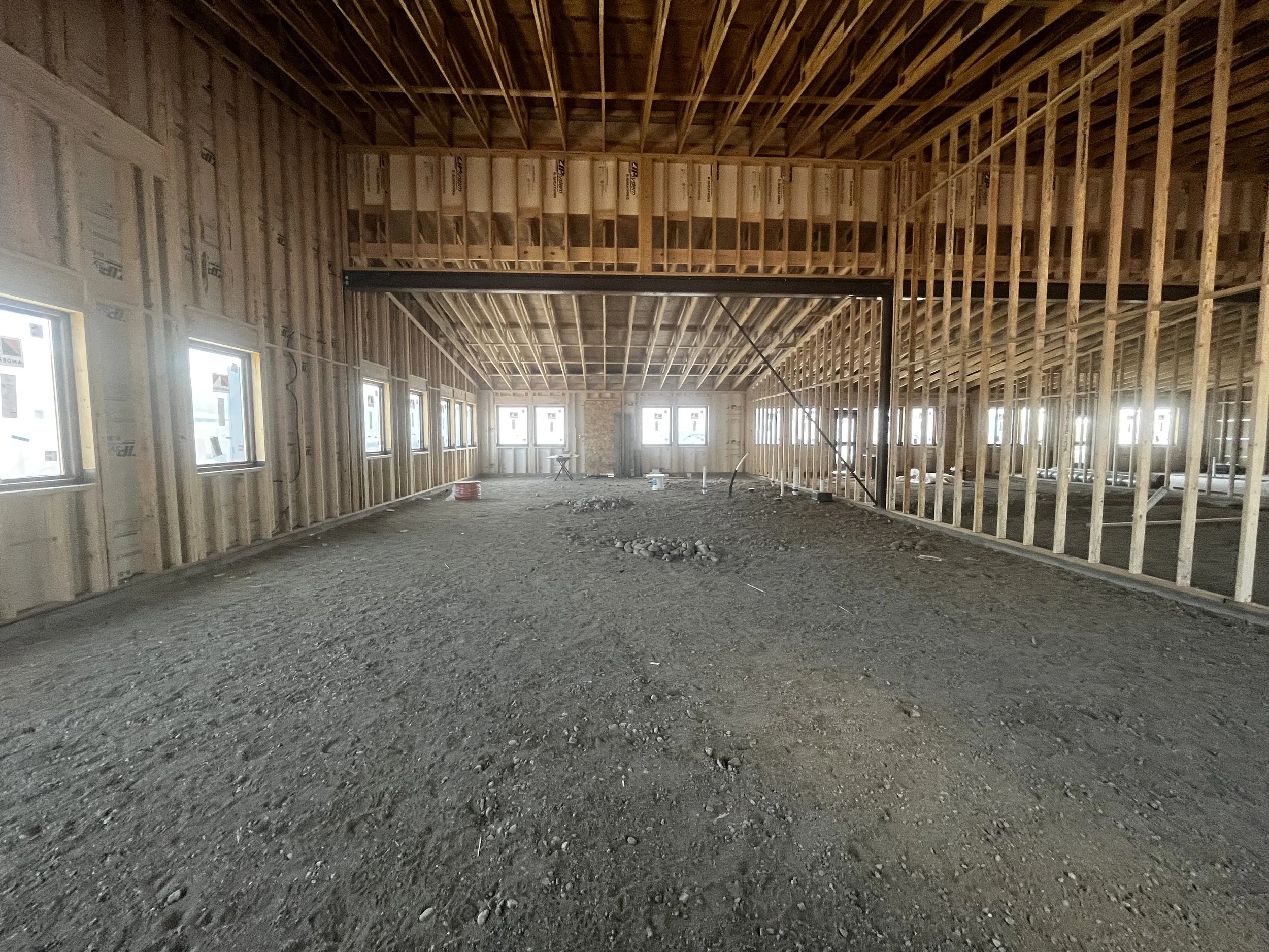 The interior of a building under construction with exposed wooden framing, multiple windows, and a rough, gravelly floor.
