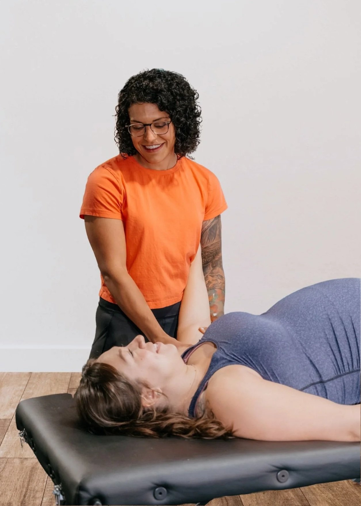 A woman lying on a massage table receiving therapy from another woman in an orange shirt, with a massage on her arm, in a room with white walls.