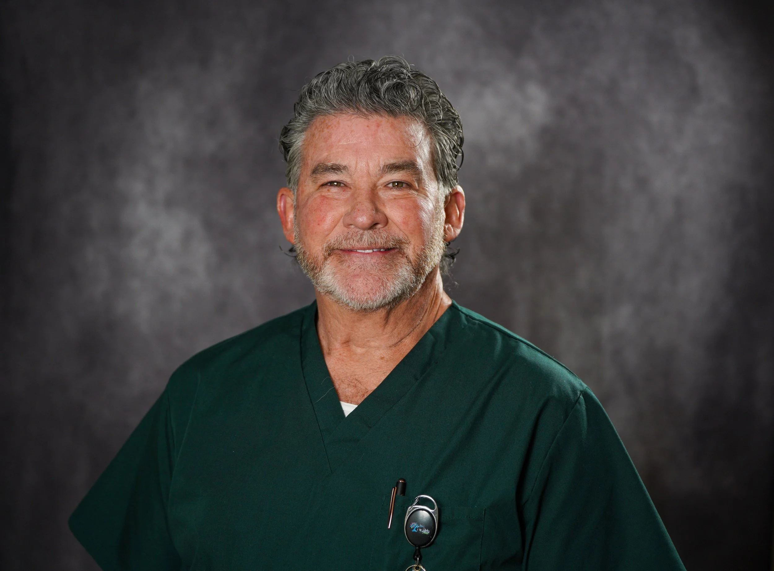 A middle-aged man with grey and black hair, wearing a dark green medical scrub uniform, smiling against a dark grey background.