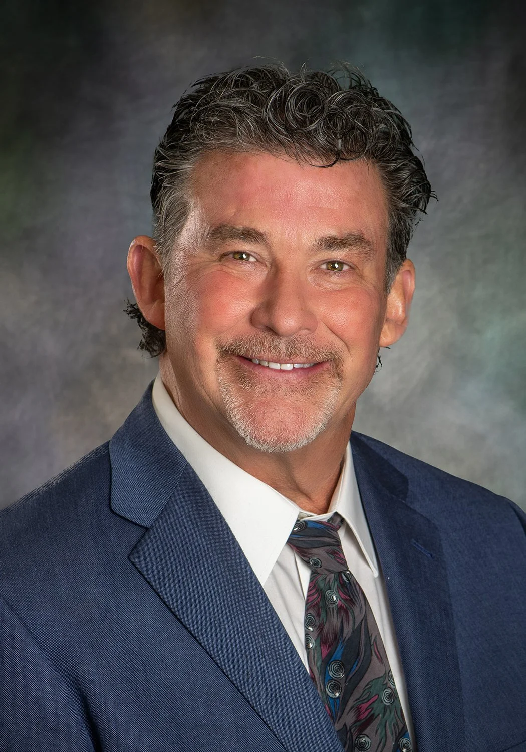 A professional headshot of a middle-aged man with gray, curly hair, a mustache, and a beard, wearing a blue suit, white shirt, and a colorful tie, smiling against a gray background.