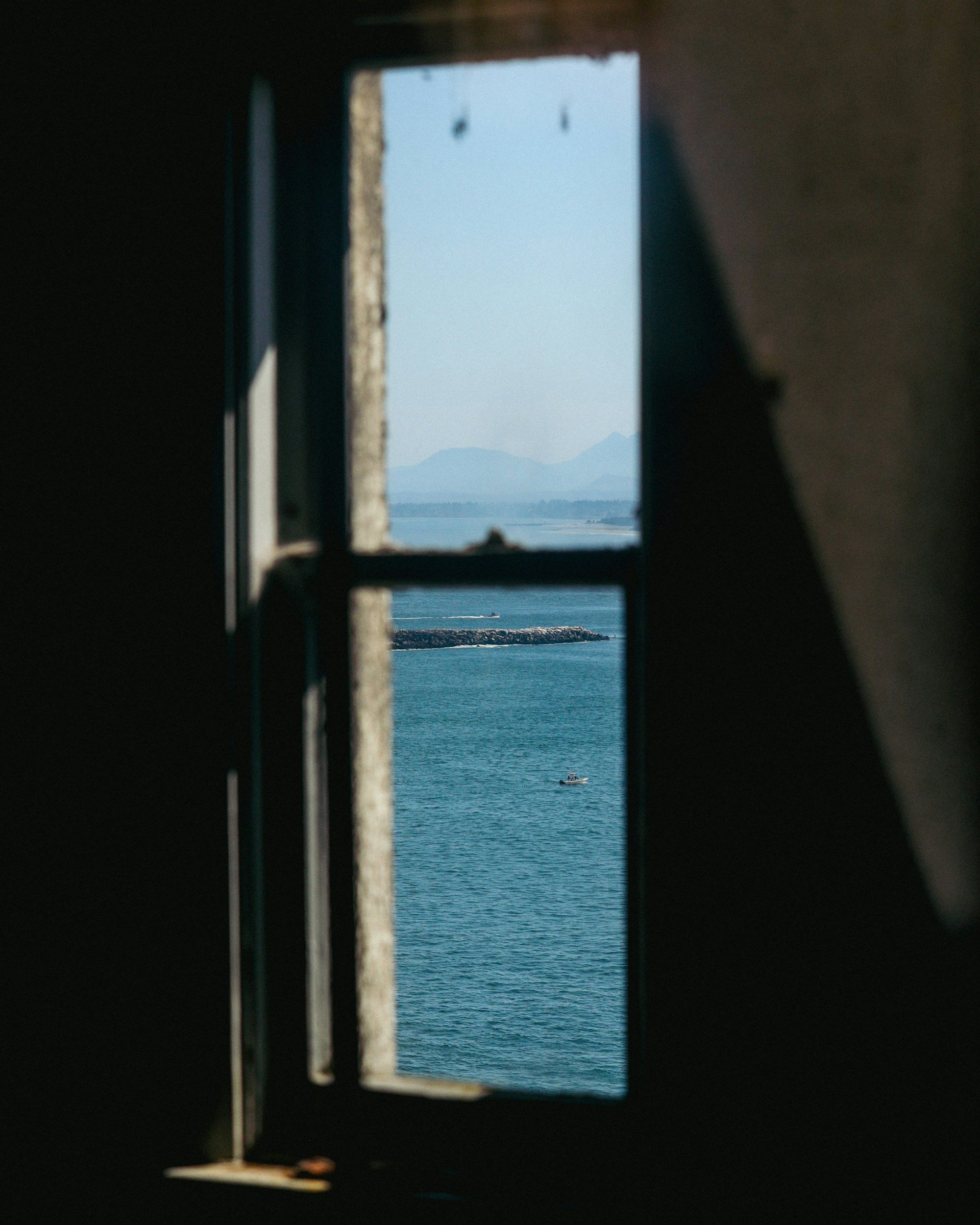 View of the ocean or sea through a small, rectangular window in a dark structure, with distant mountains on the horizon and a boat on the water.