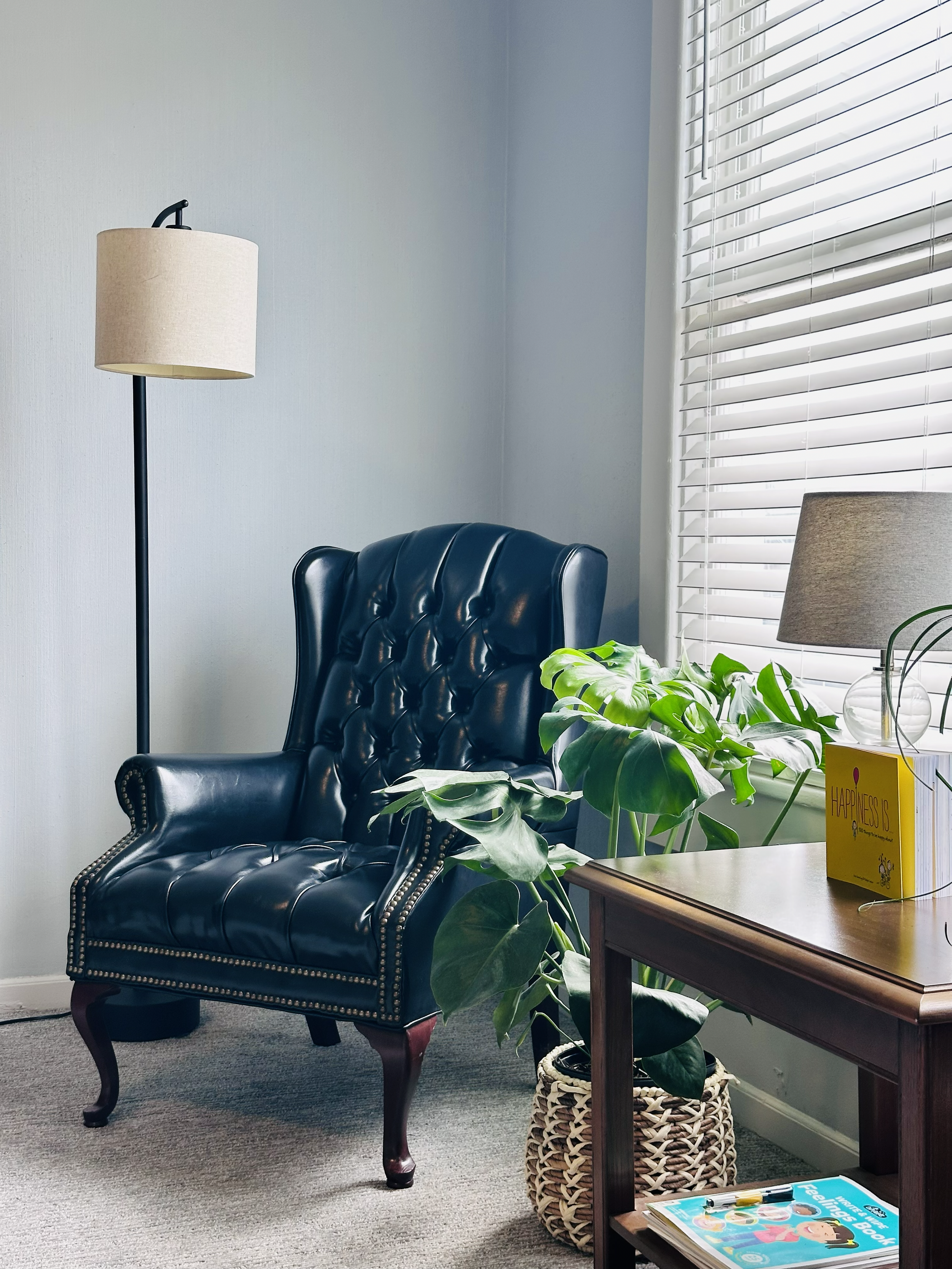 A cozy corner featuring a black leather armchair with nailhead trim, a wooden side table with a yellow happiness book and colorful markers, a large green houseplant in a woven basket, a beige table lamp with a fabric shade, and a tall floor lamp with