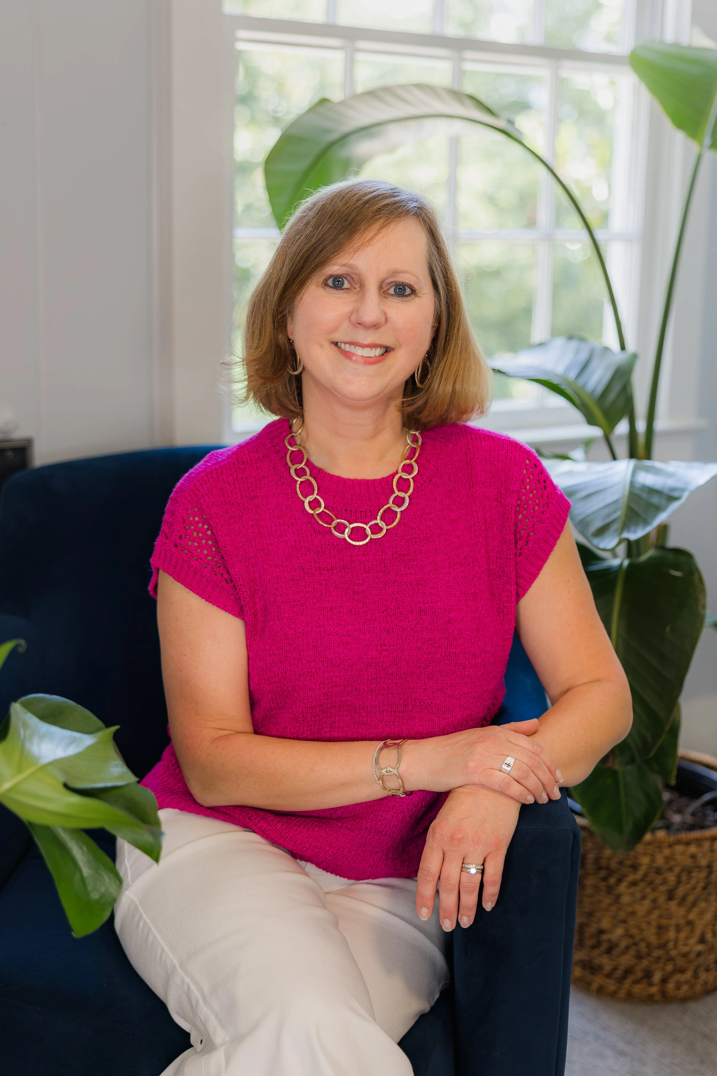 A woman with shoulder-length brown hair, wearing a pink top, white pants, and jewelry, sitting on a dark blue chair indoors near a large window and green houseplants.