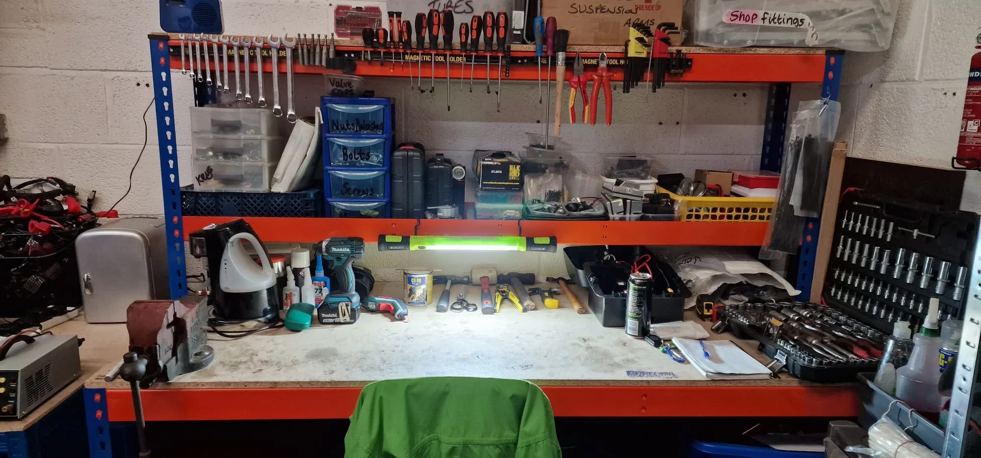 A well-organized workshop workbench with tools, parts, and storage containers. The workbench has tools hanging above, a level, and various tools and supplies laid out on the surface. Shelves and bins hold smaller items and parts.