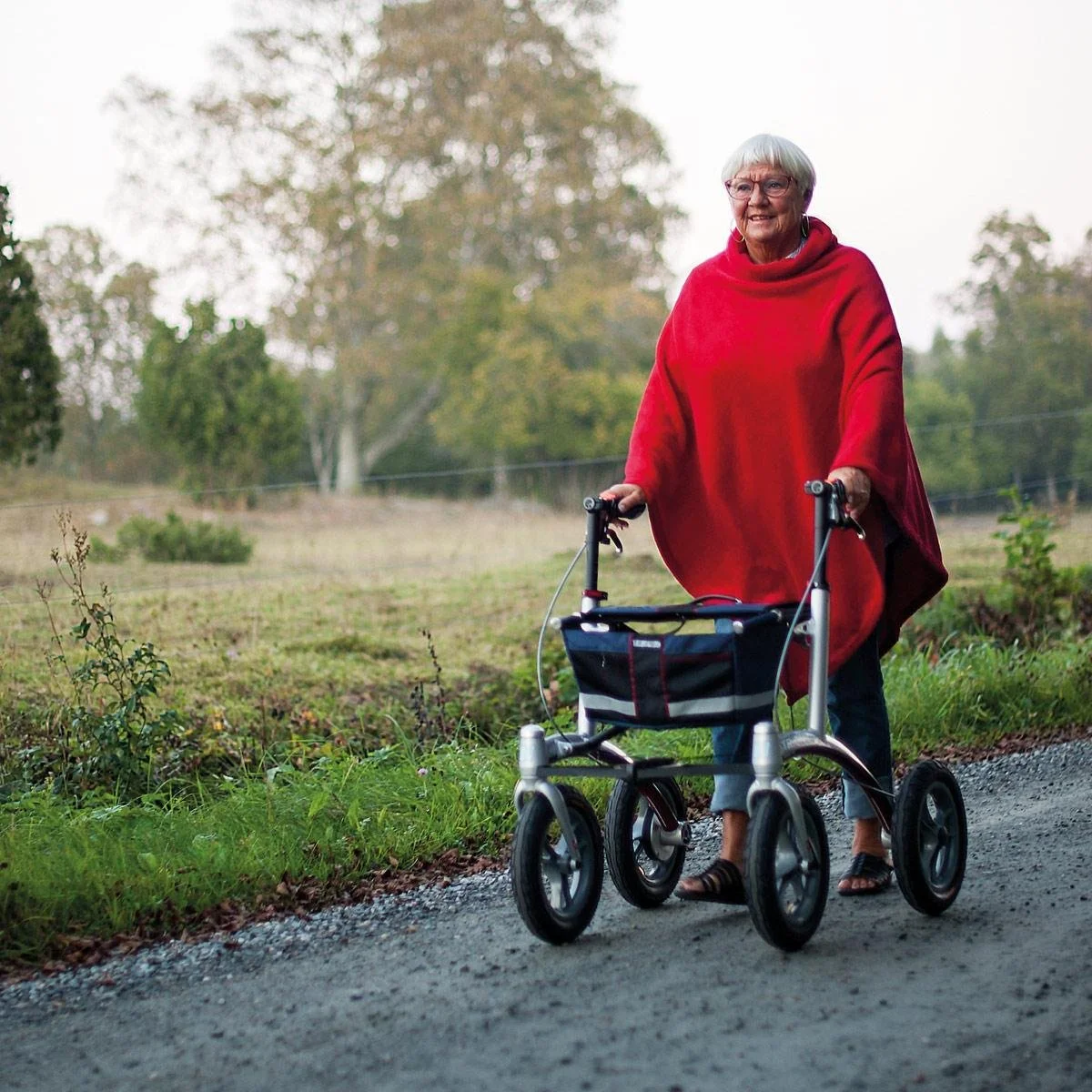 An elderly woman with gray hair and glasses, wearing a red poncho, smiling as she walks outdoors with a walker on a gravel path, surrounded by trees and grass.