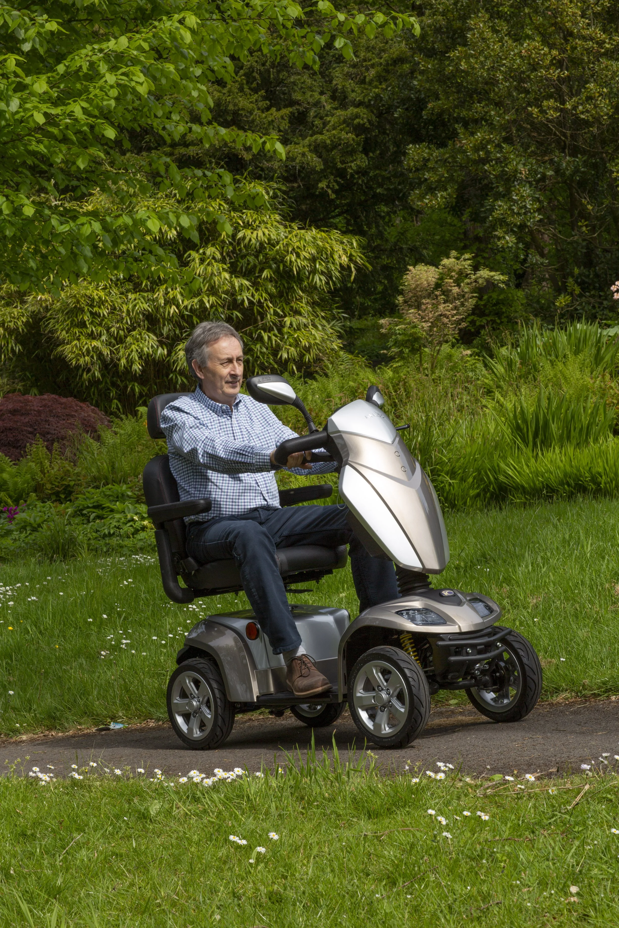 An elderly man with gray hair and a checkered shirt riding a mobility scooter on a grassy area with flowers. The background features lush green trees and shrubs.