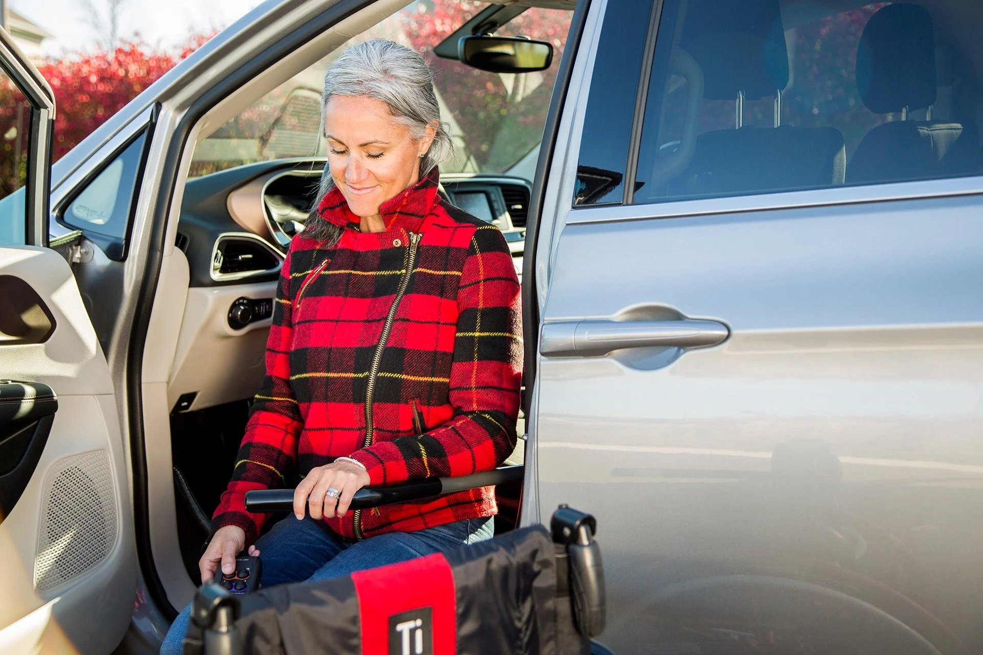 An older woman with gray hair, wearing a red and black plaid jacket, gets into a silver vehicle using a wheelchair. She is smiling and looking down, with a background of trees with fall foliage.