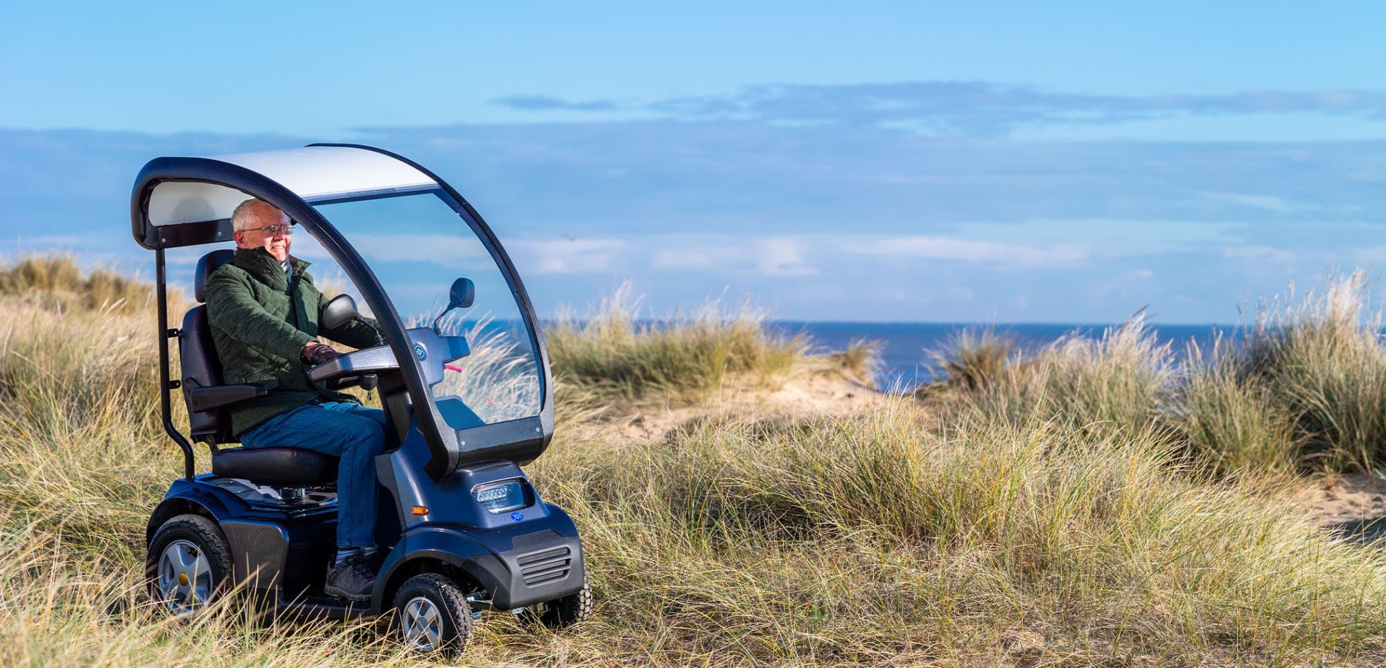 A man in a mobility scooter driving through a grassy coastal area with sand dunes and the ocean in the background under a partly cloudy sky.