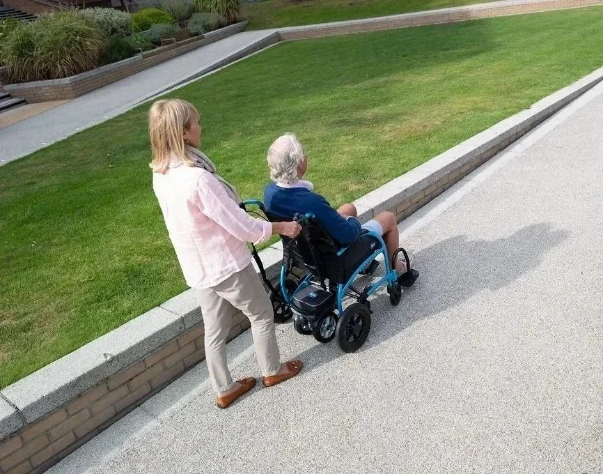 A woman pushing an elderly person in a wheelchair along a sidewalk beside a grassy area at what appears to be a park or garden.