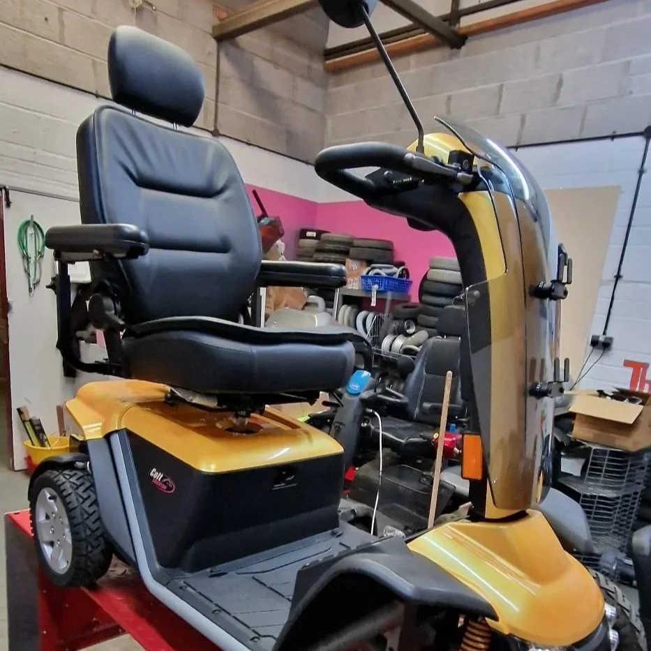 A yellow mobility scooter with a black seat and armrests, parked inside a storage or workshop space with tools and supplies around.