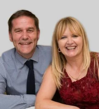 A smiling middle-aged man in a light blue dress shirt and dark tie sitting beside a smiling middle-aged woman with blonde hair wearing a red lace top.