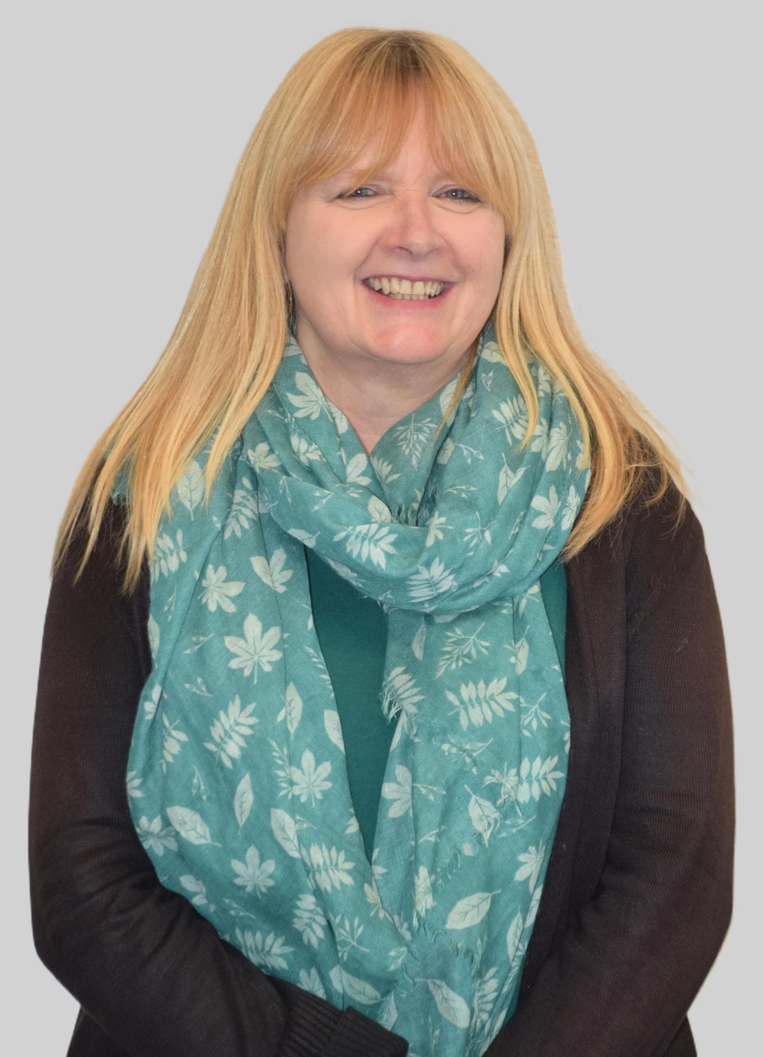 A woman with long blonde hair, wearing a teal scarf with white floral patterns and a dark brown jacket, sitting against a plain light gray background, smiling.