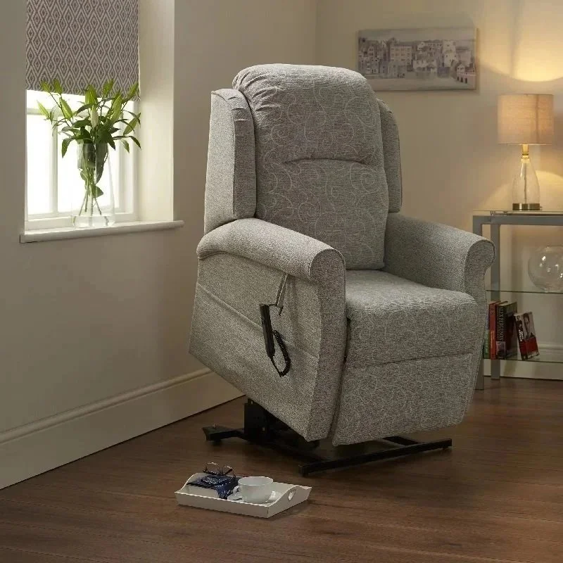 A gray recliner chair in a living room, next to a window with a potted plant, a side table with a lamp, and a tray with a teacup on the floor.