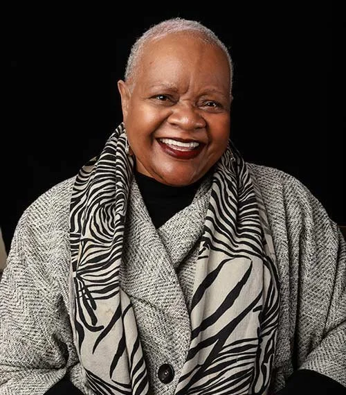 Portrait of an elderly woman smiling, wearing a patterned scarf and a gray sweater, with a black background.