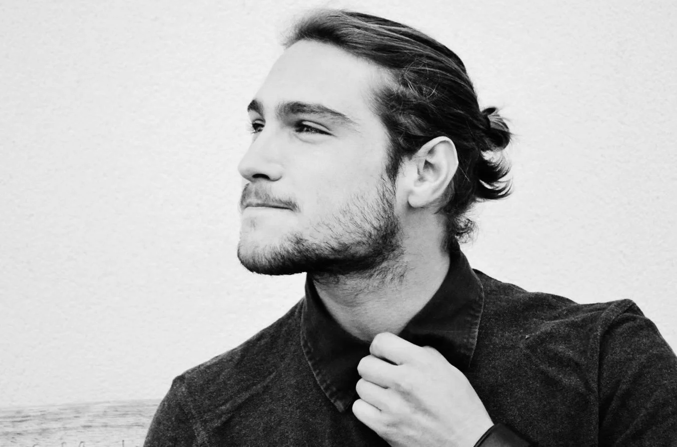 Black and white portrait of a man with long hair tied back, looking to the side, touching his collar with one hand, against a plain wall background.
