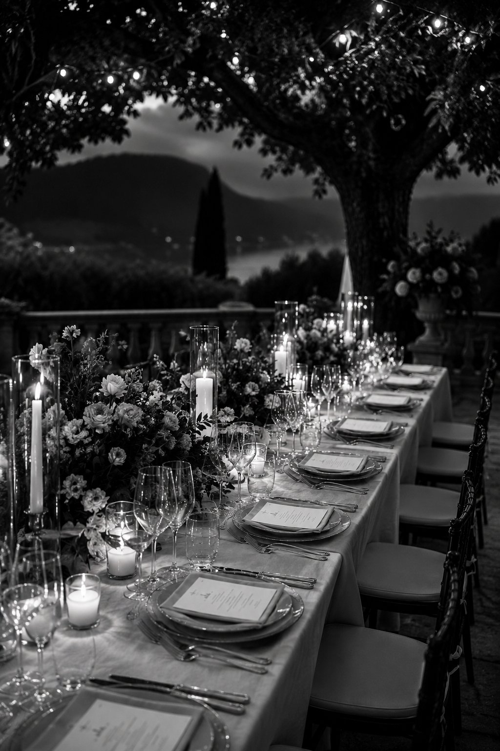 Black and white outdoor dinner table set with candles, flowers, and wine glasses under a tree with string lights, overlooking a landscape with mountains and water.