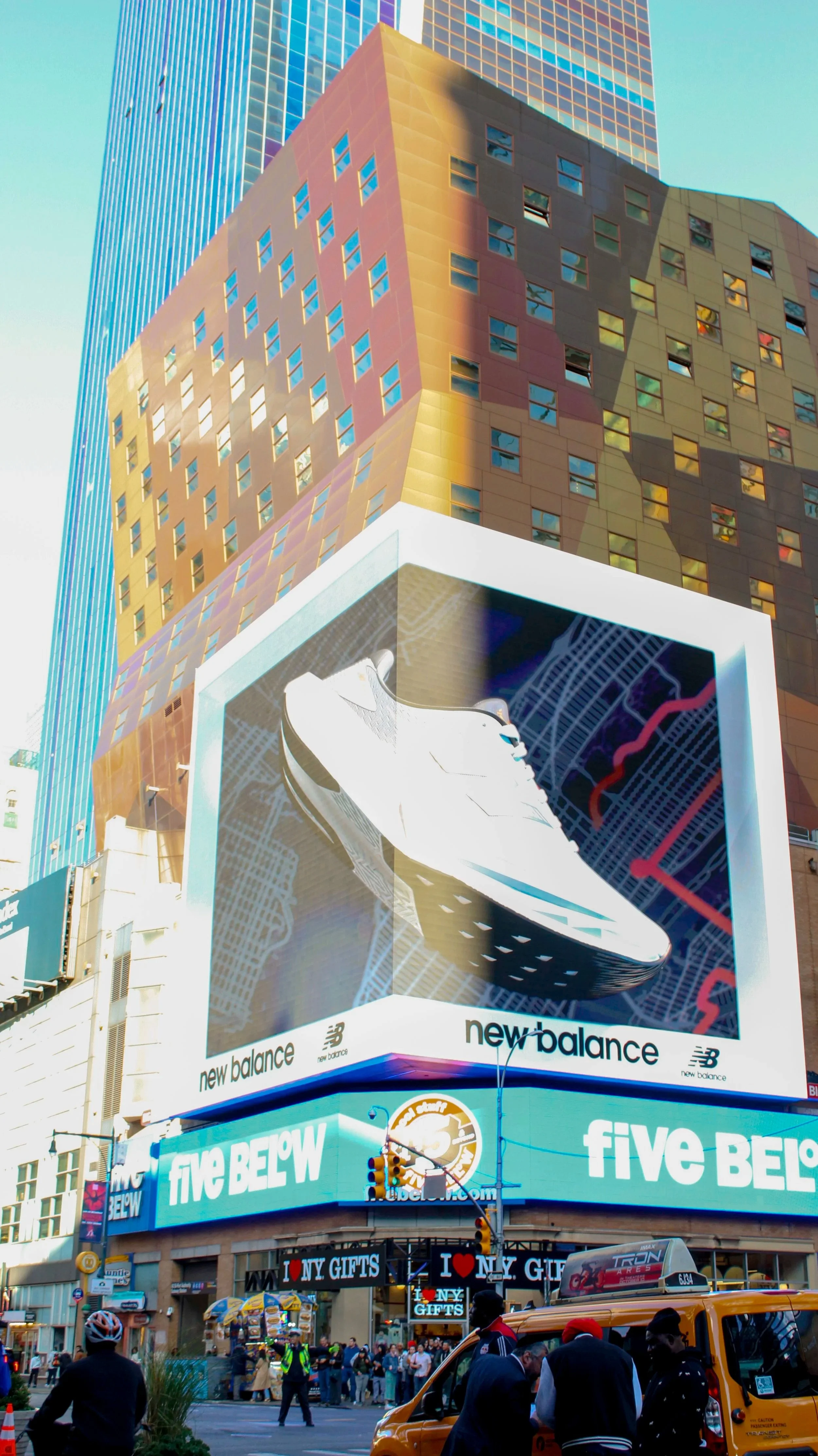 A digital billboard featuring a New Balance sneaker, with the background of tall buildings and a crowd of pedestrians and yellow taxis on a busy city street.