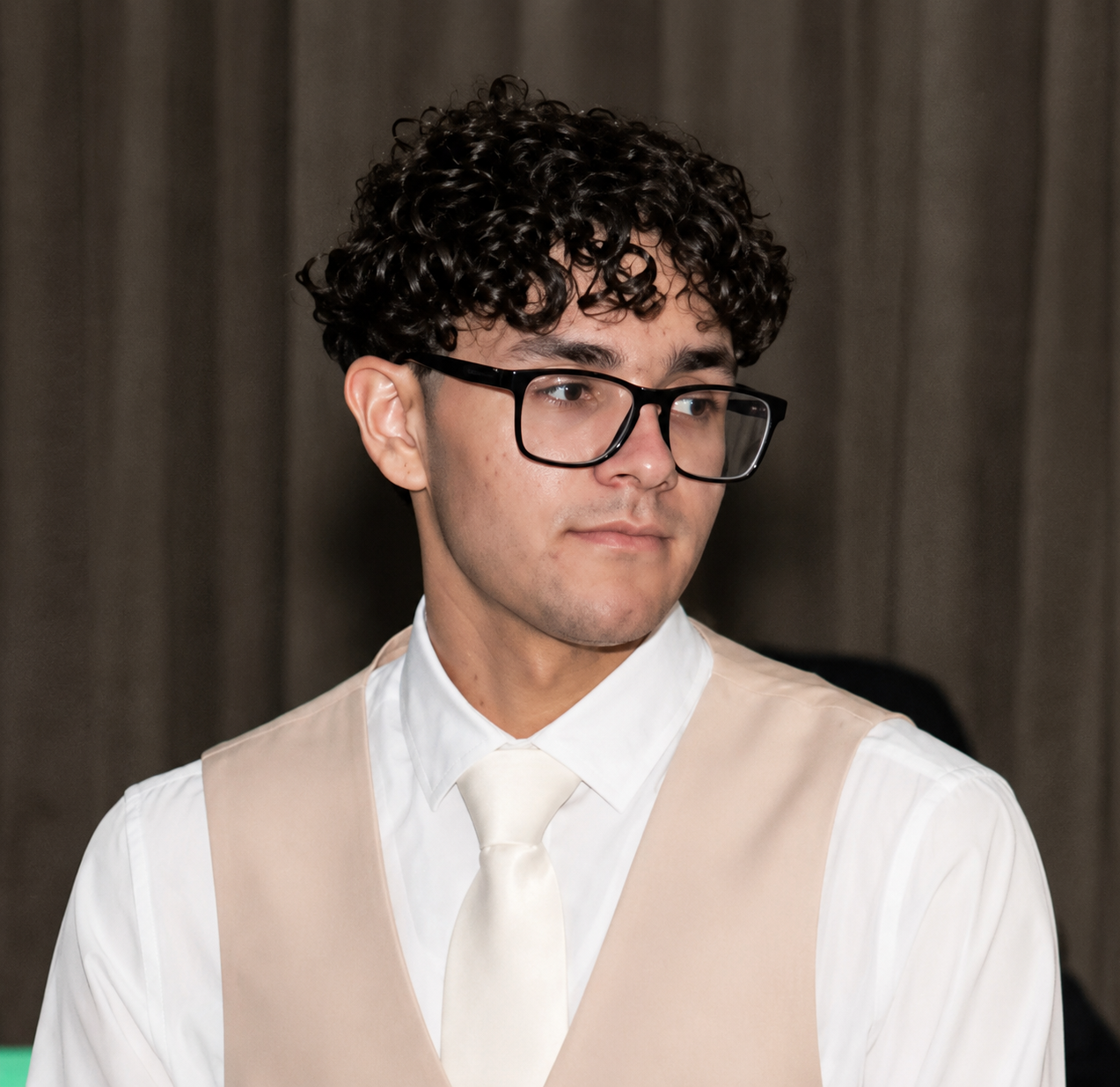 A young man with curly dark hair and glasses, wearing a white shirt and beige vest with a cream tie, looking slightly to the side, in front of a gray curtain.