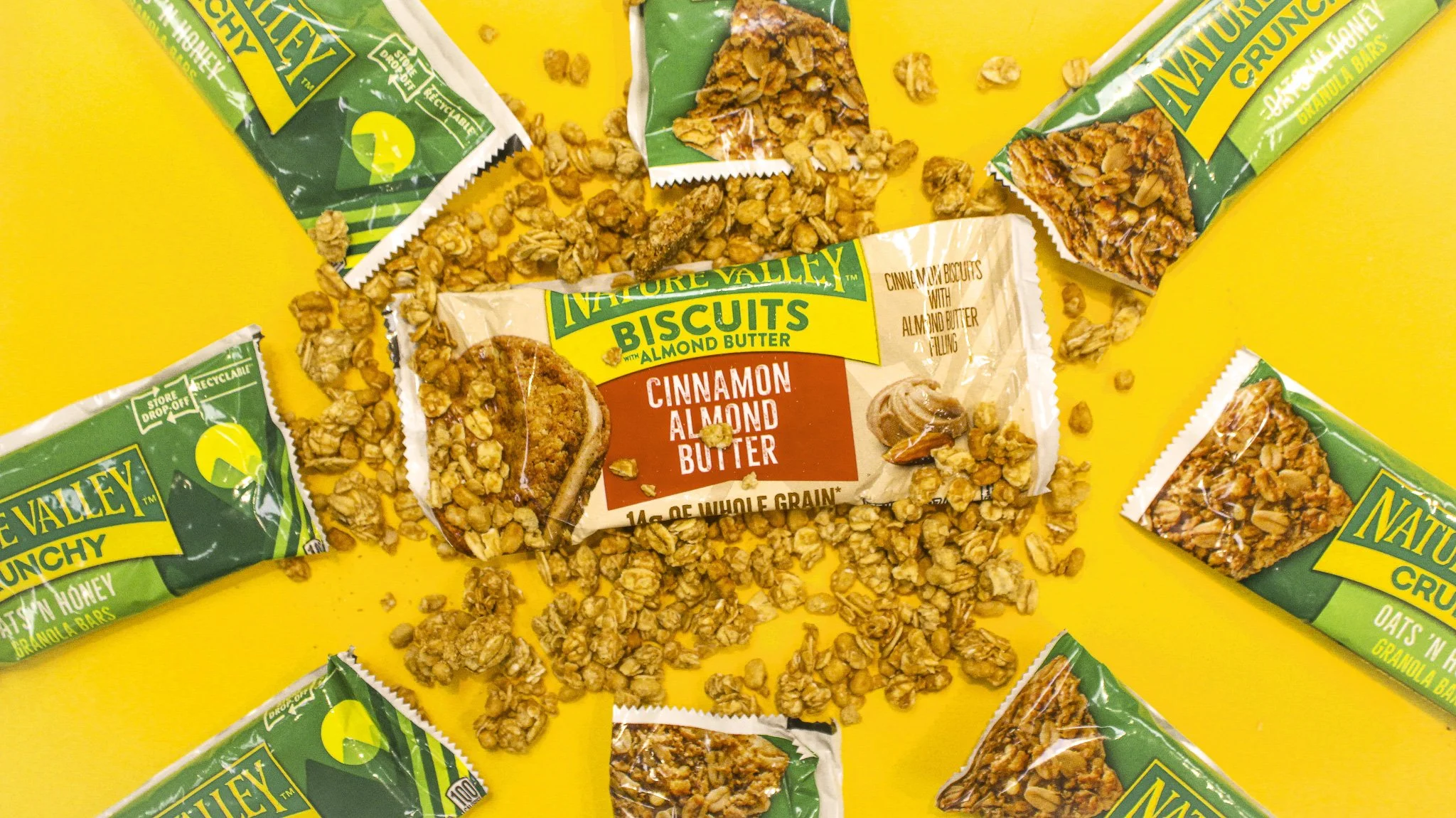 Unopened Nature Valley granola bars with almond butter flavor, surrounded by granola pieces on a yellow background.