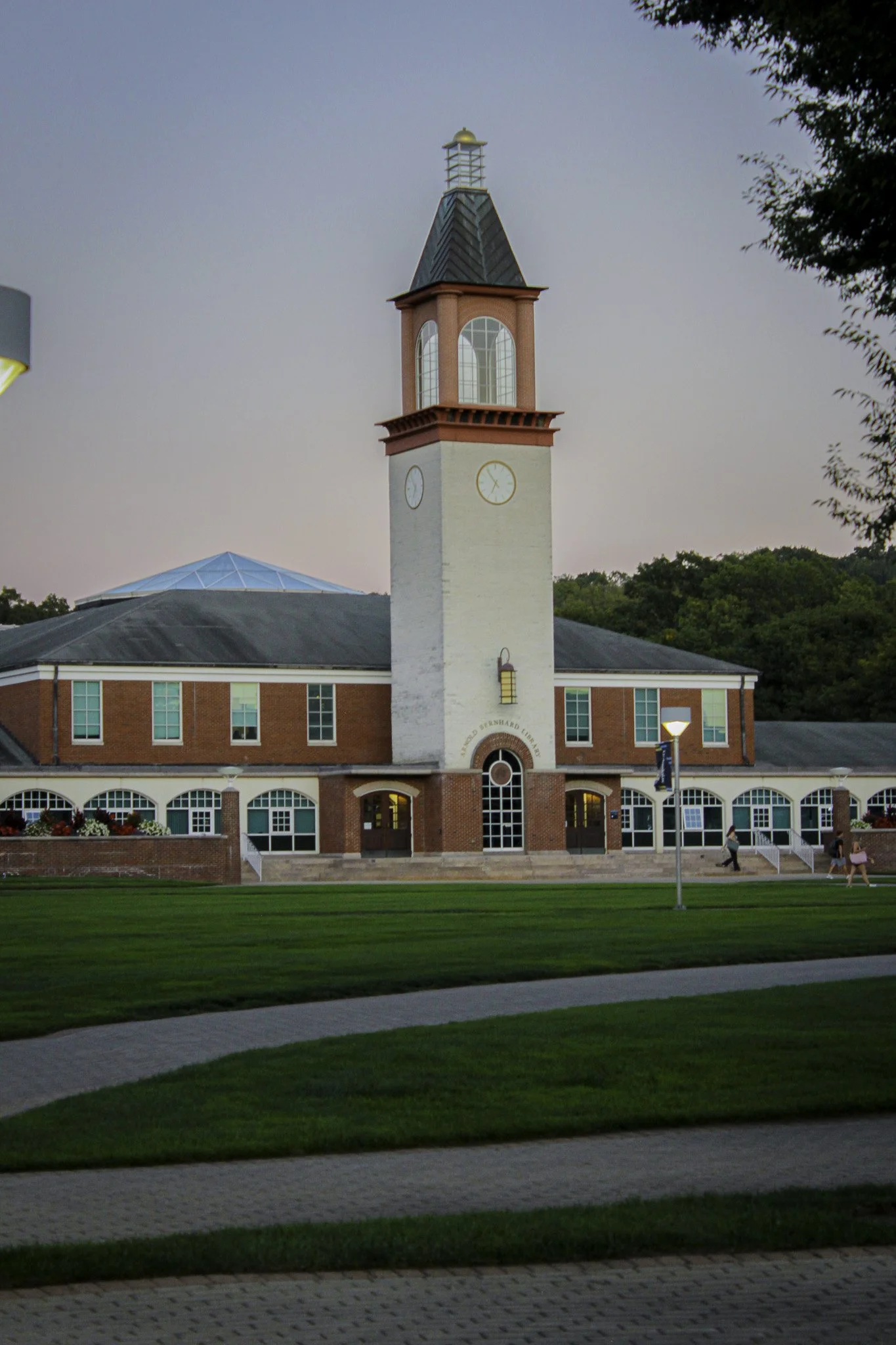 A red brick building with white trim and a clock tower, situated in a park-like setting with a well-maintained lawn, trees, and a few pedestrians walking along the pathway.