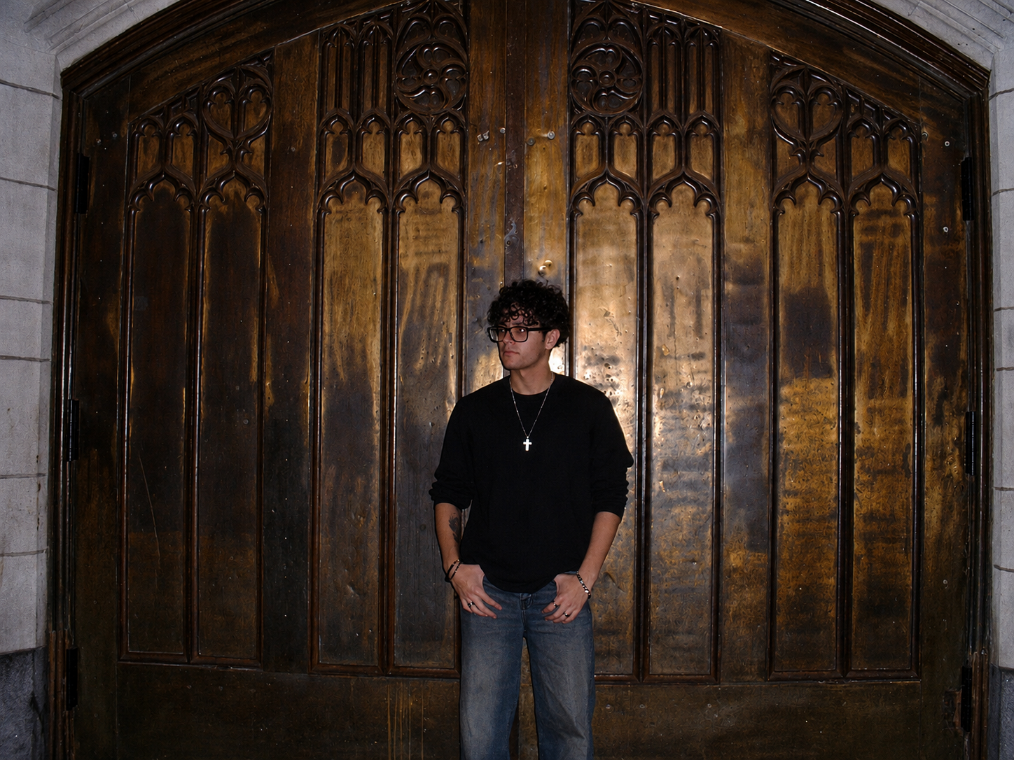 A young man with curly hair, glasses, and a cross necklace standing in front of a large, ornate wooden door.