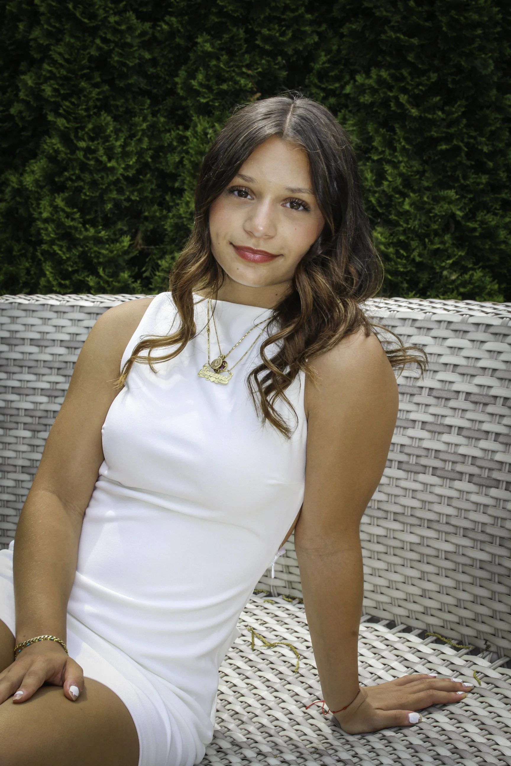 Young woman with long brunette hair sitting outdoors on wicker furniture, wearing a white sleeveless dress, with greenery in the background.