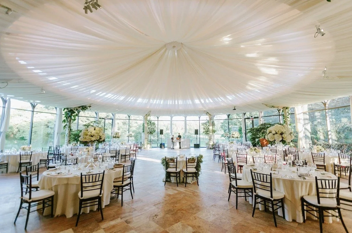 Elegant indoor wedding reception with round tables covered in white tablecloths, decorated with floral centerpieces, under a draped white ceiling with natural light coming through large windows.