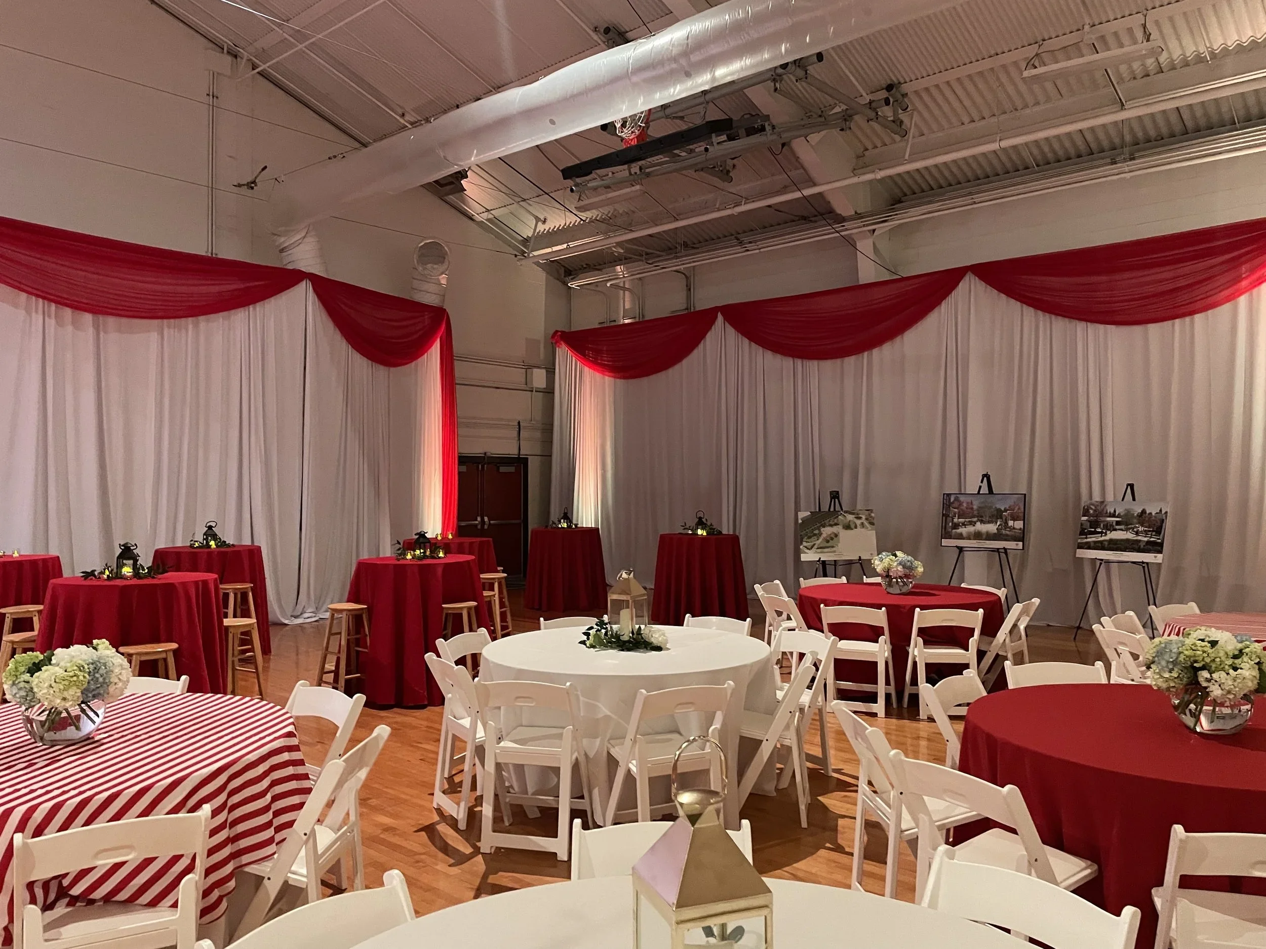 Event banquet hall decorated with red and white drapery, round tables with red and striped tablecloths, floral centerpieces, and white folding chairs, set up for a celebration or reception.