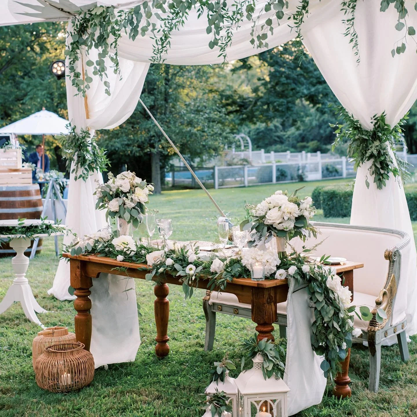 Elegant outdoor wedding or event setup with a wooden table decorated with white flowers and greenery, a white sofa, lanterns, and draped white curtains with greenery accents under a canopy, surrounded by trees and a white fence in the background.