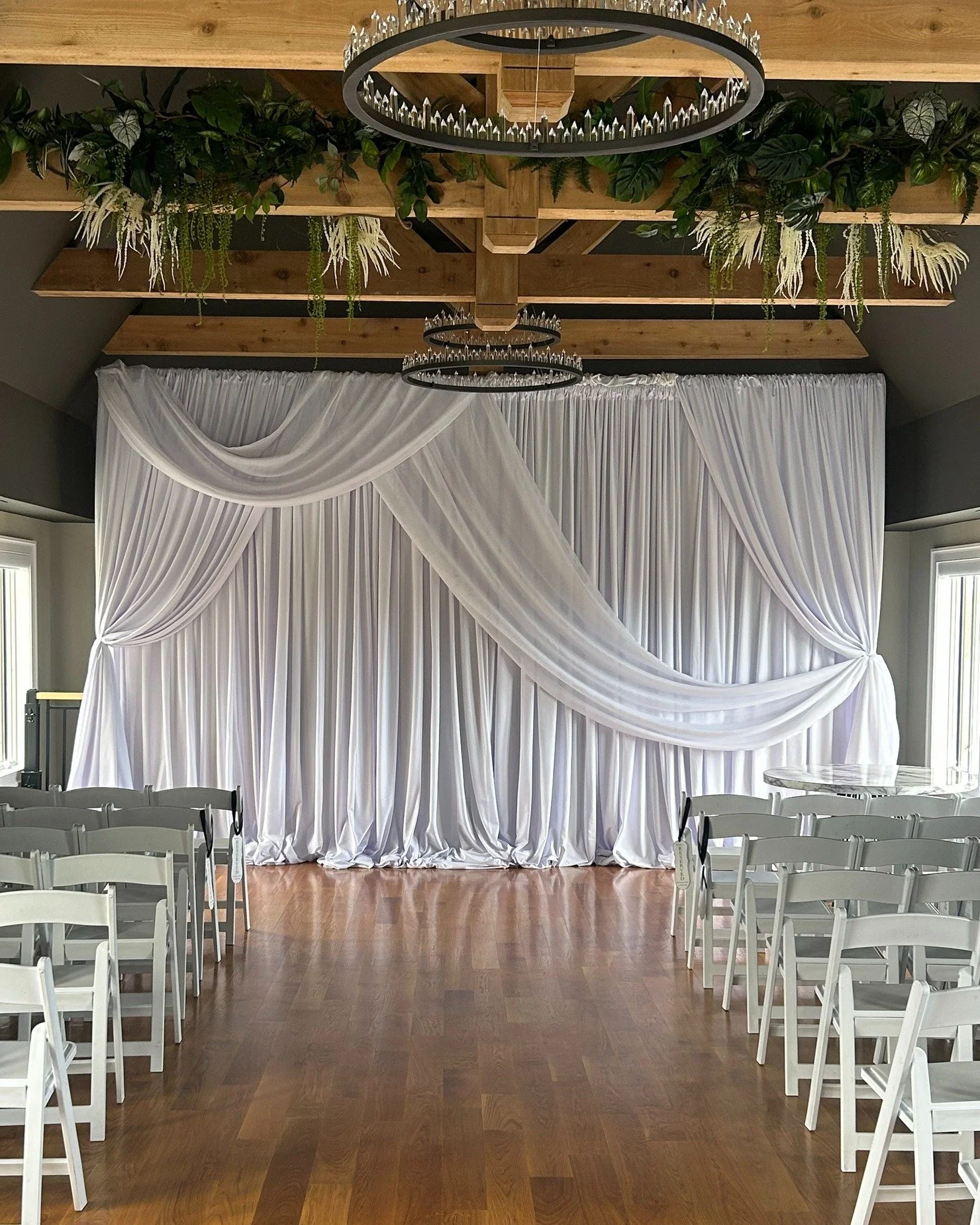 Indoor wedding ceremony setup with white chairs facing a stage with white drapes and a flower-and-greenery backdrop, and a circular chandelier hanging from a wooden ceiling.